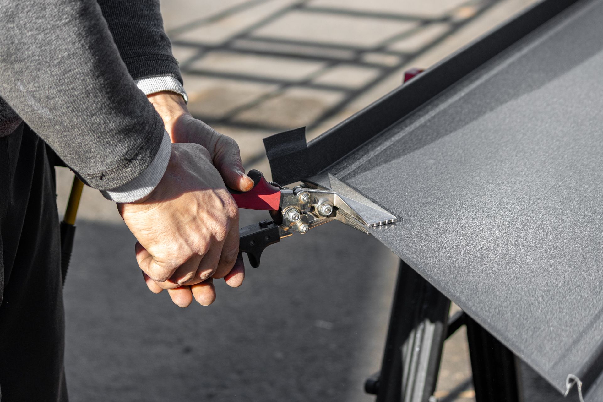 Person using metal shears to cut a piece of gray sheet metal on a work table.