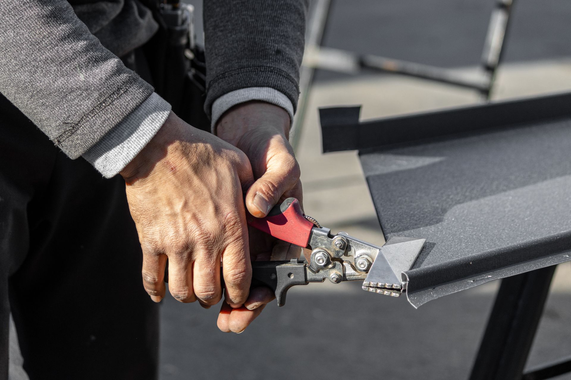 Person using red-handled metal shears to cut a piece of dark metal roofing outside.