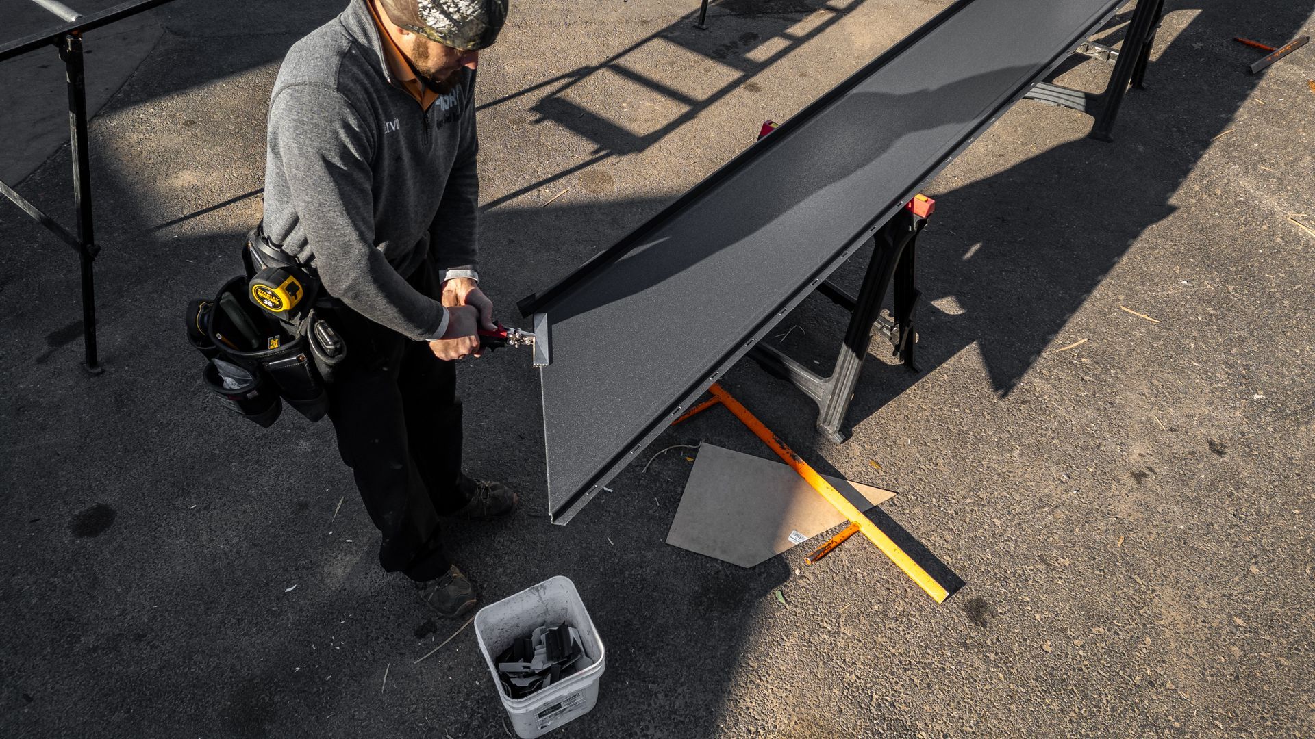 A worker in gray sweatshirt uses tools to work on a dark panel on sawhorses outdoors.
