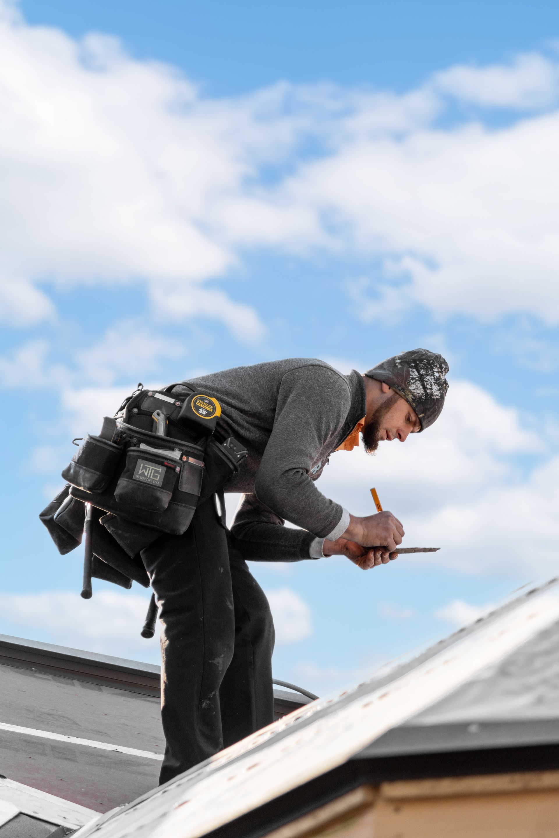 Roofer on a roof, wearing tool belt, writing on a clipboard against a cloudy sky.
