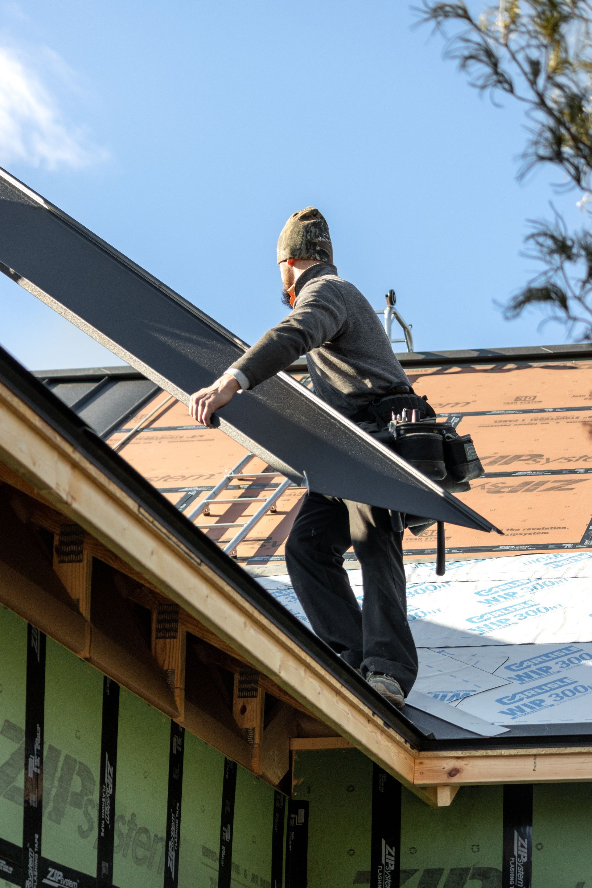 Roofer carrying a dark metal sheet on a rooftop, wearing a hat and tool belt.