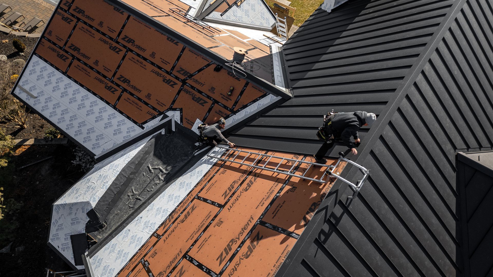 Roofers working on a multi-level roof with dark metal and orange underlayment, outdoors.