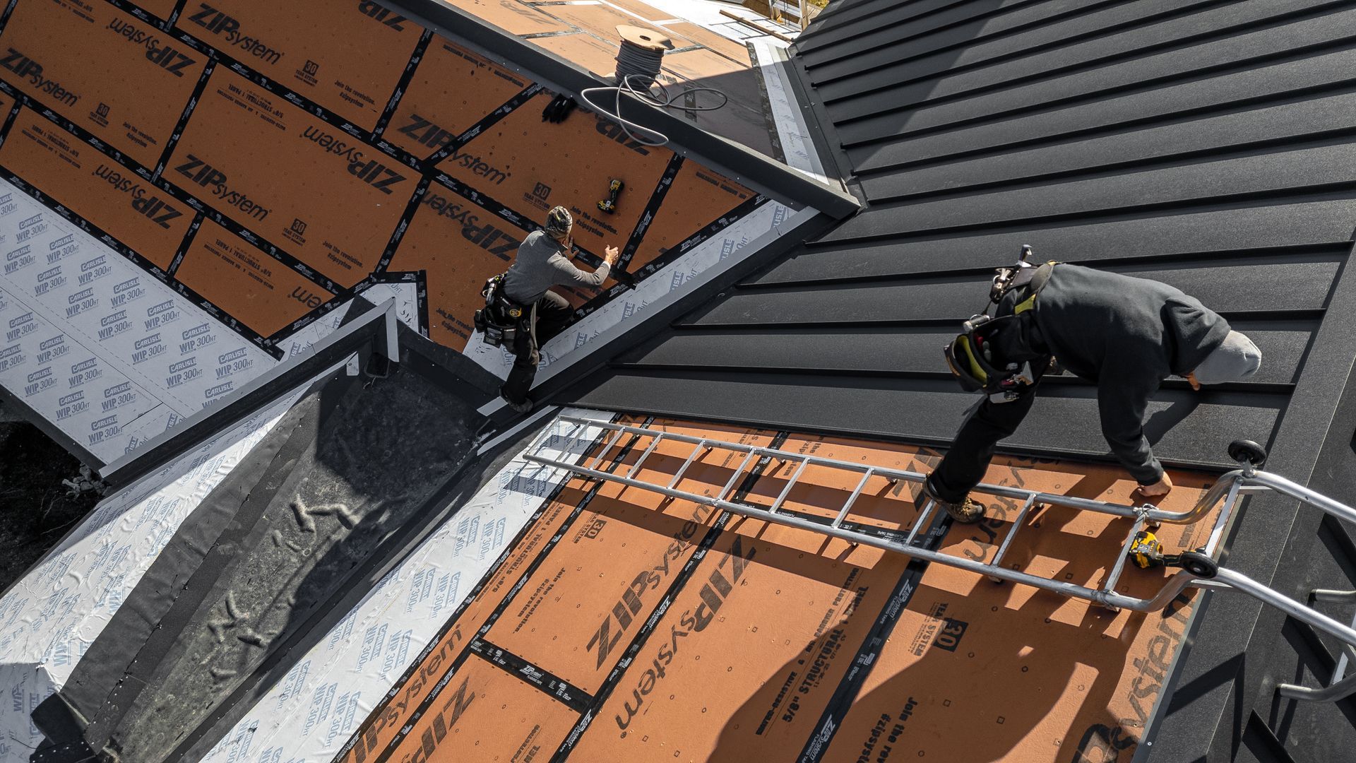 Roofers working on a roof, applying sheathing. One worker uses a ladder. Brown and black tones.
