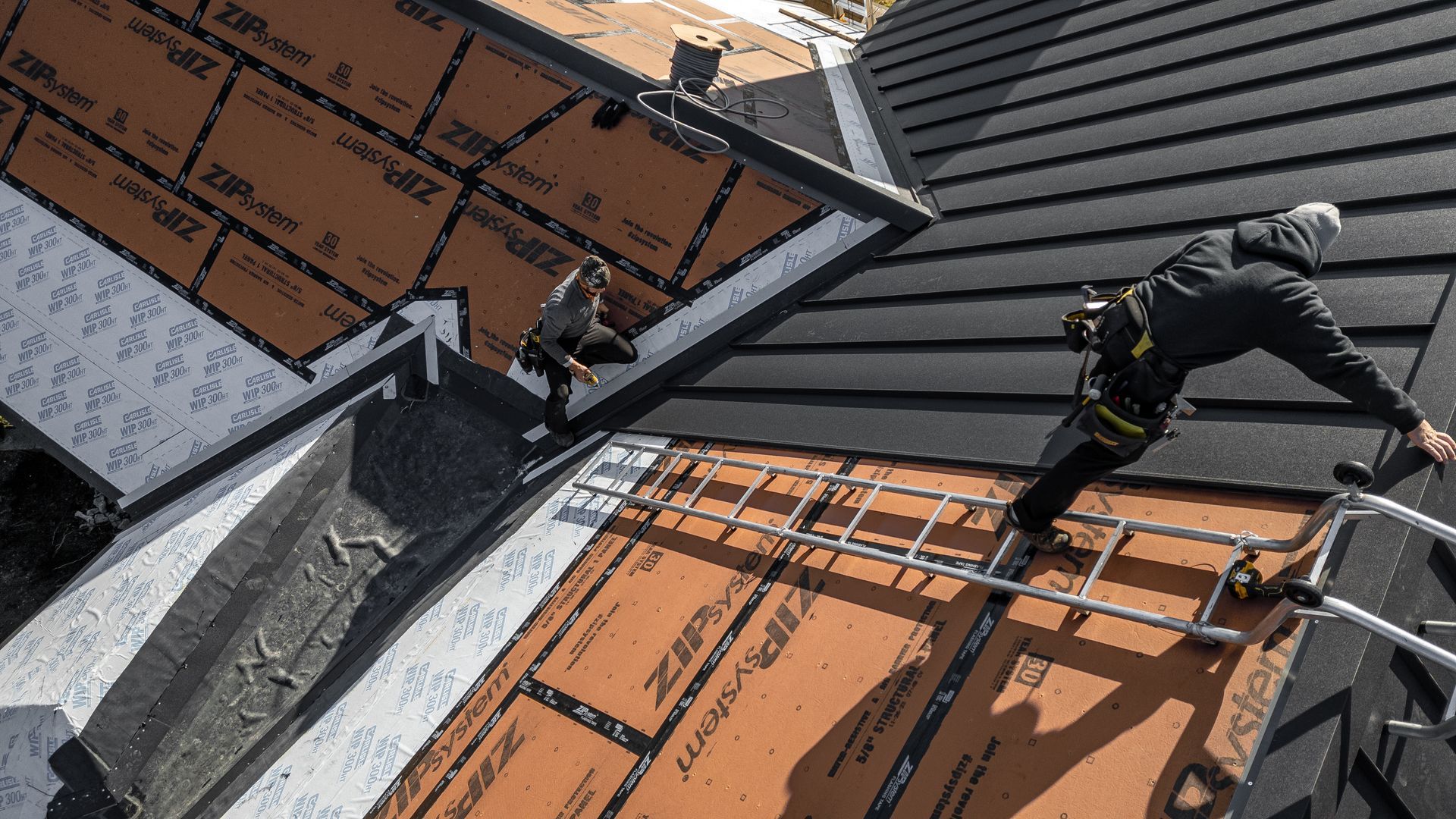 Roofer with harness working on dark shingle roof and brown sheathing by Midtech Standing Seam