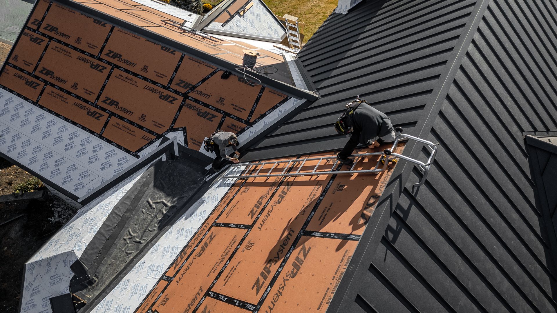 Two workers installing roofing material on a house. Black metal roof and tan underlayment.