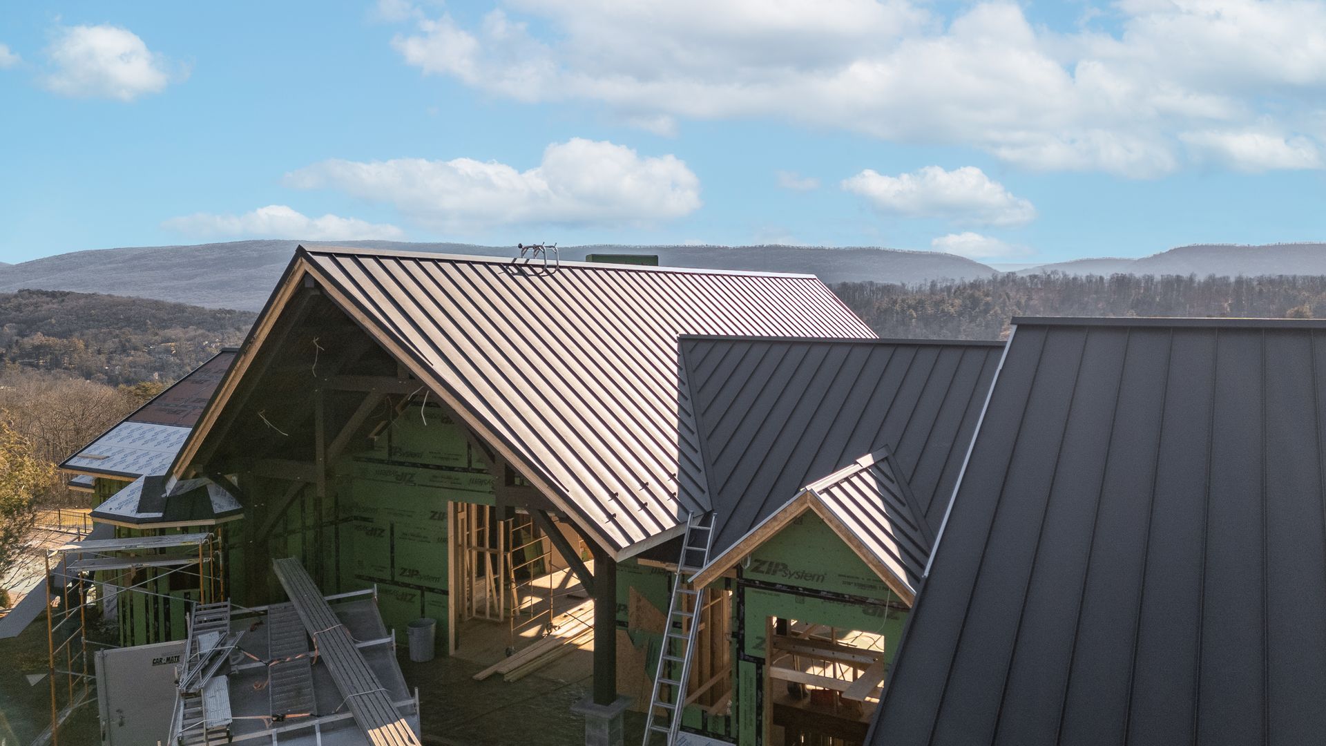 A partially constructed house with dark metal roof, wood framing, and green wrap, set in a mountainous landscape.