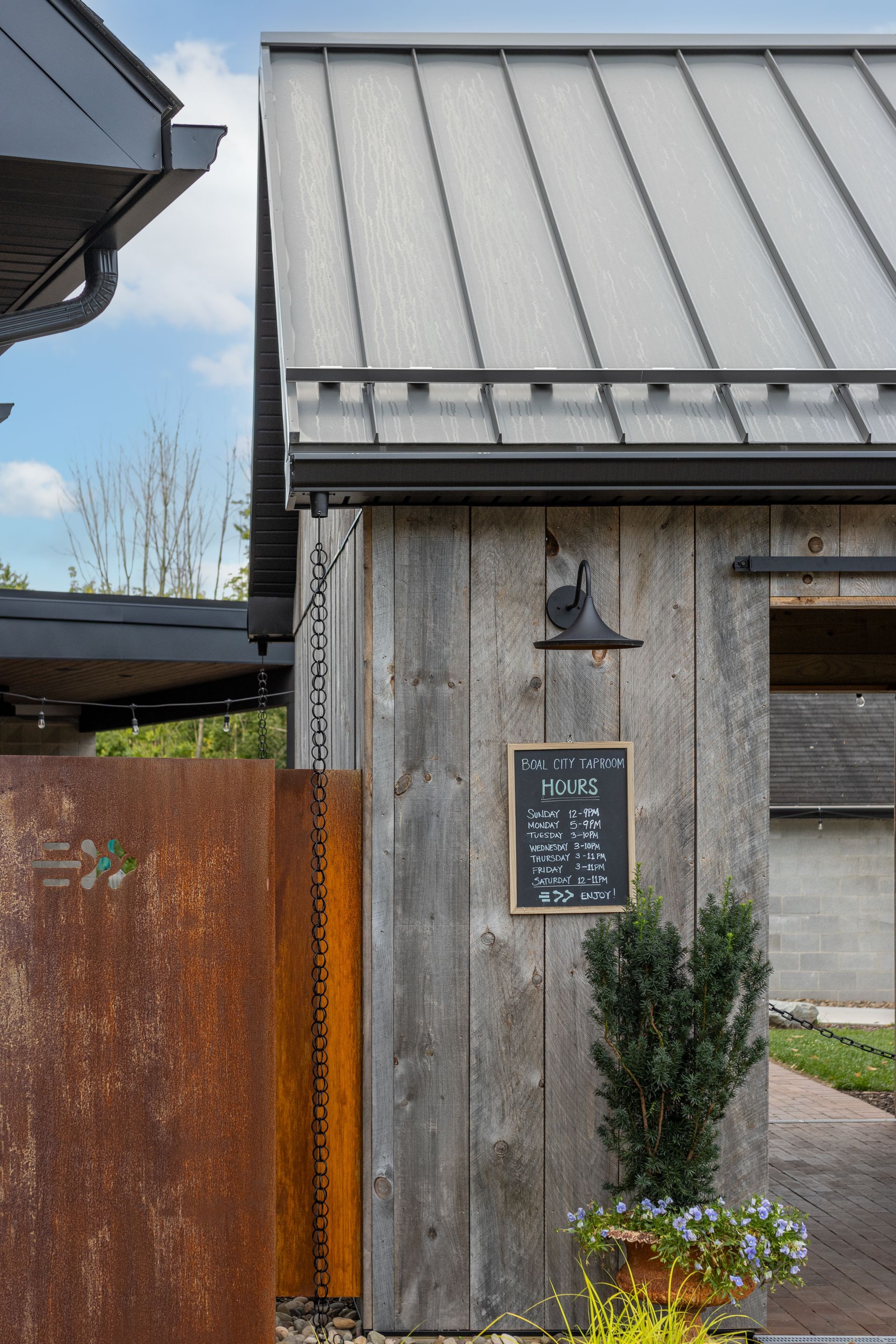 Rustic building exterior with weathered wood, metal roof, sign, and potted plants.
