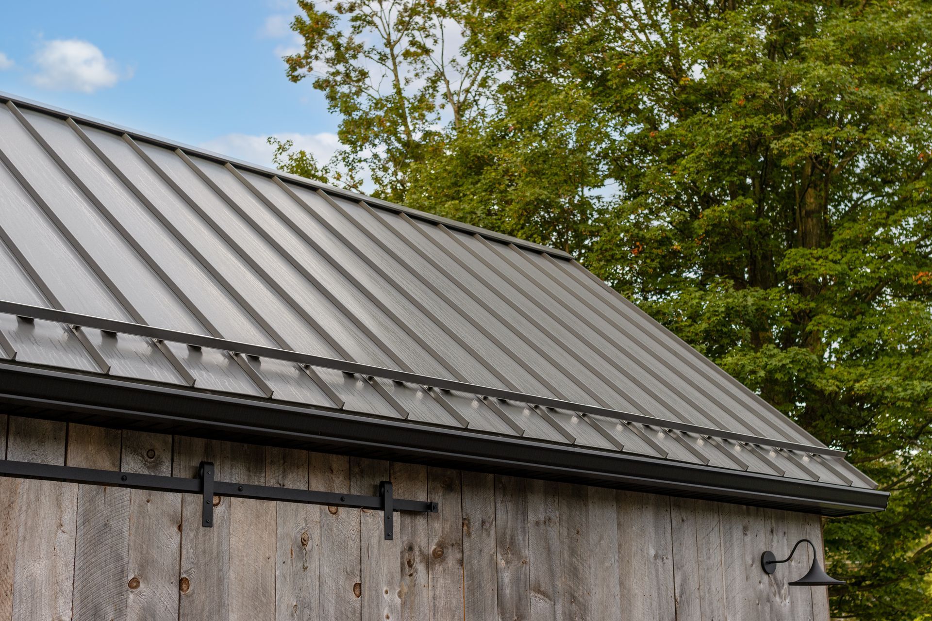 Gray metal roof on a weathered wooden barn with a black trim, set against a green tree and blue sky.