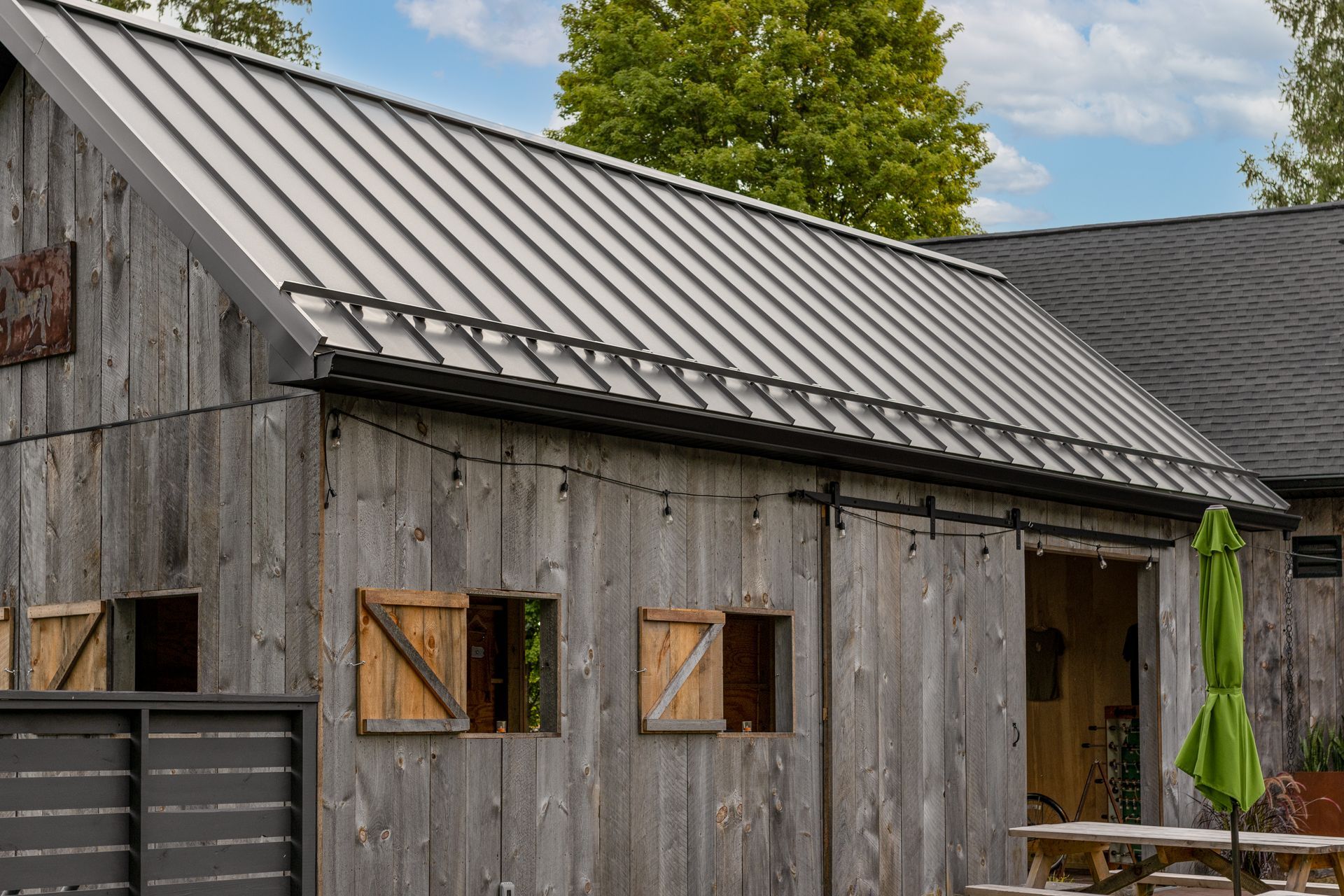 Wooden barn with a metal roof, shutters, and open doorway. Green umbrella stands nearby.