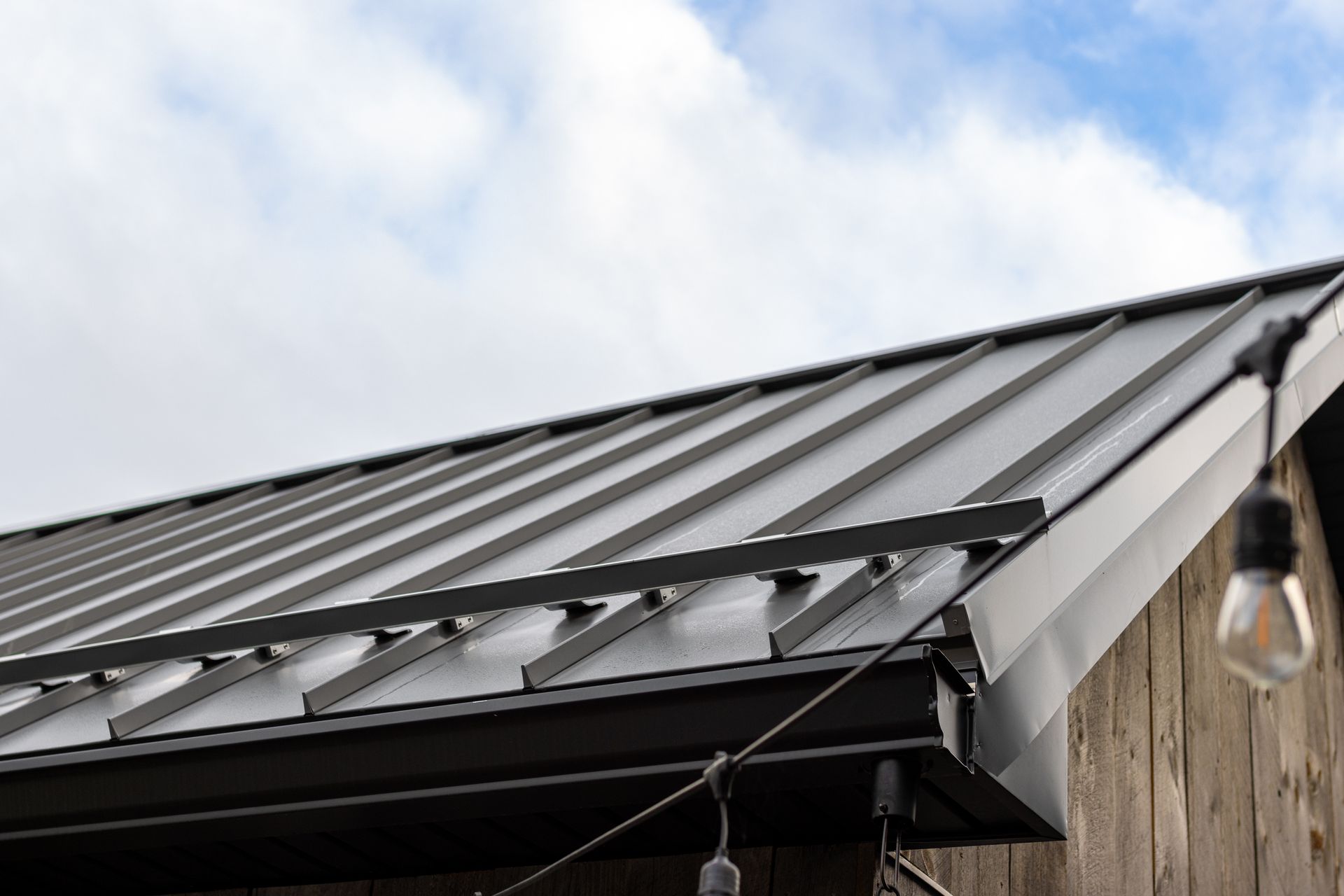 Black metal roof on a building, with a cloudy sky background and string lights.