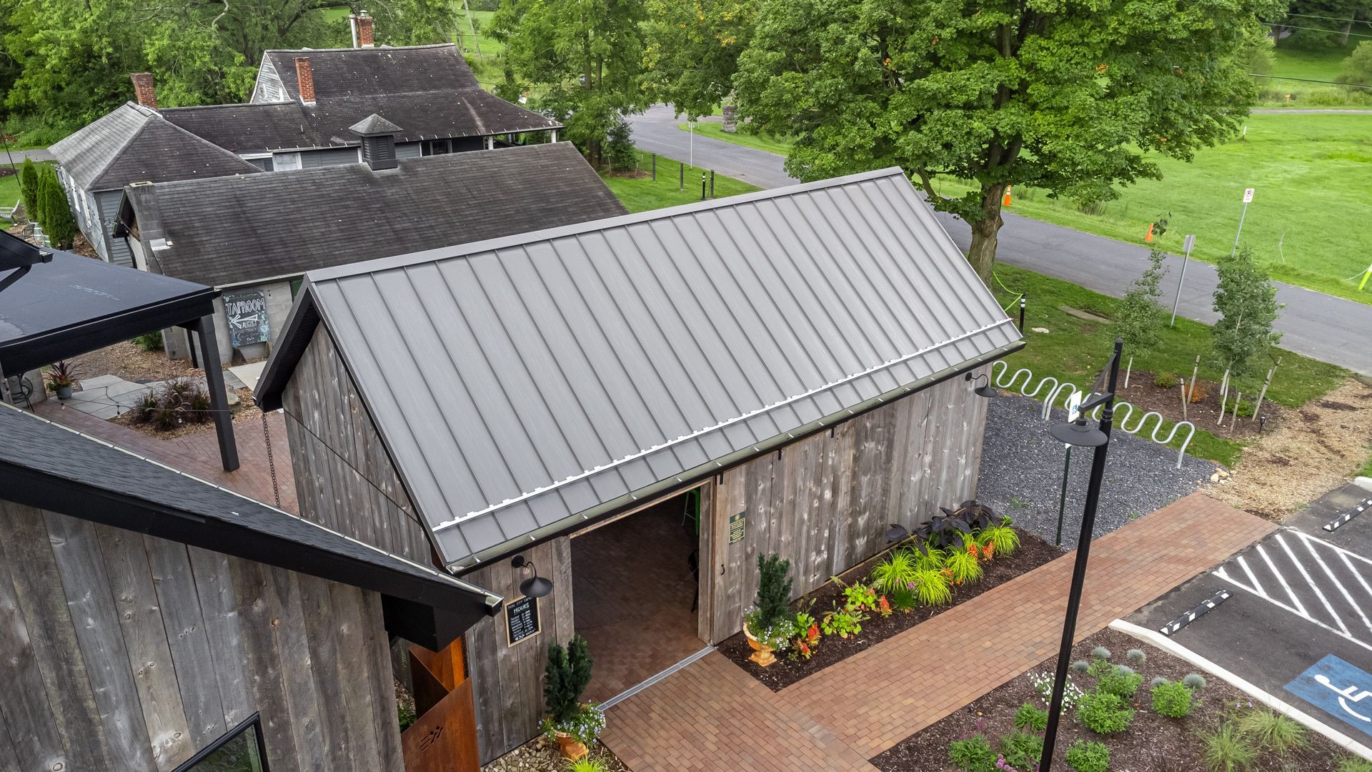 Wooden barn with a metal roof, next to a brick walkway, with a building in the background.