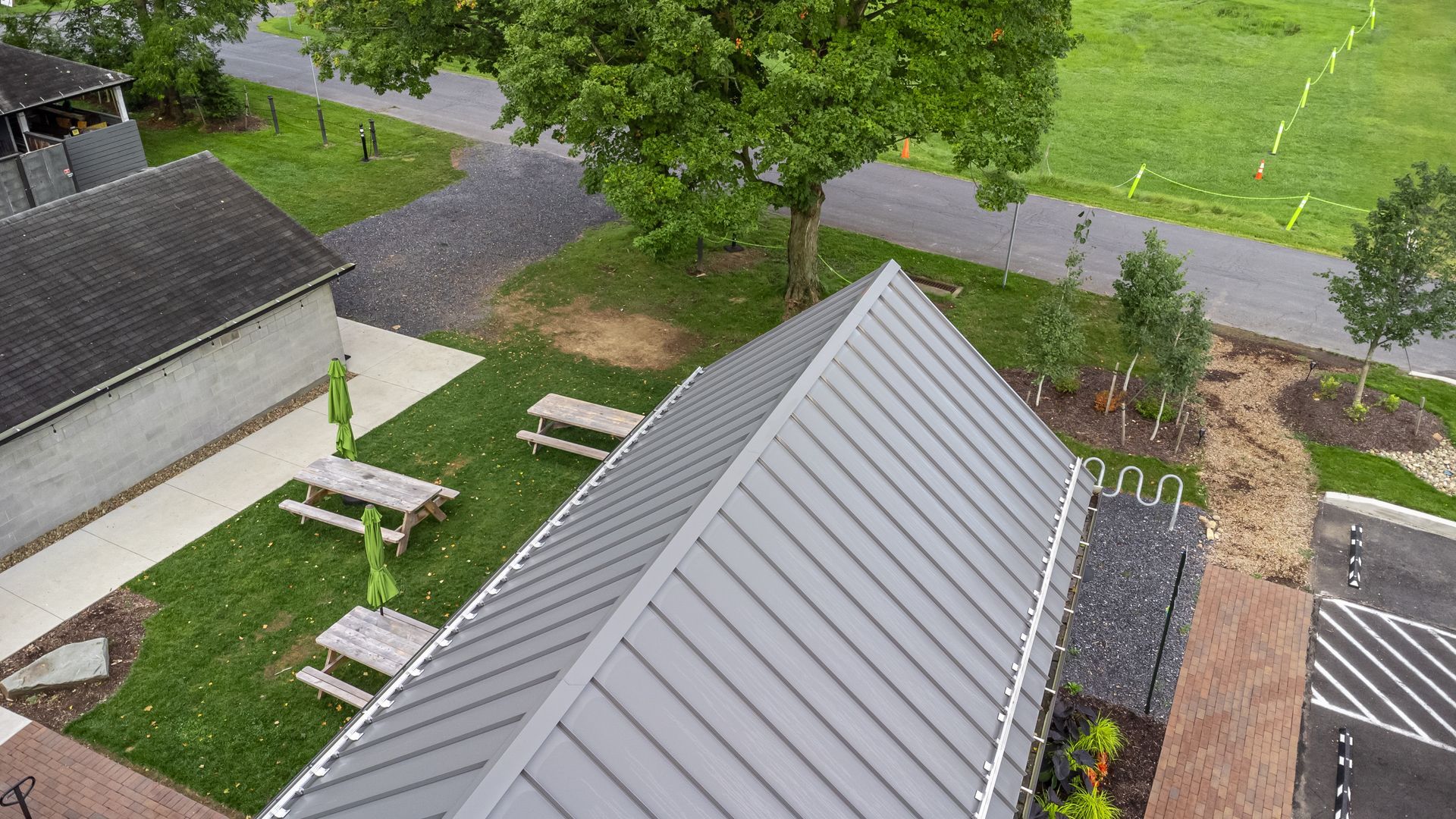 Overhead view of building with gray metal roof, picnic tables on grass, next to a road with trees.