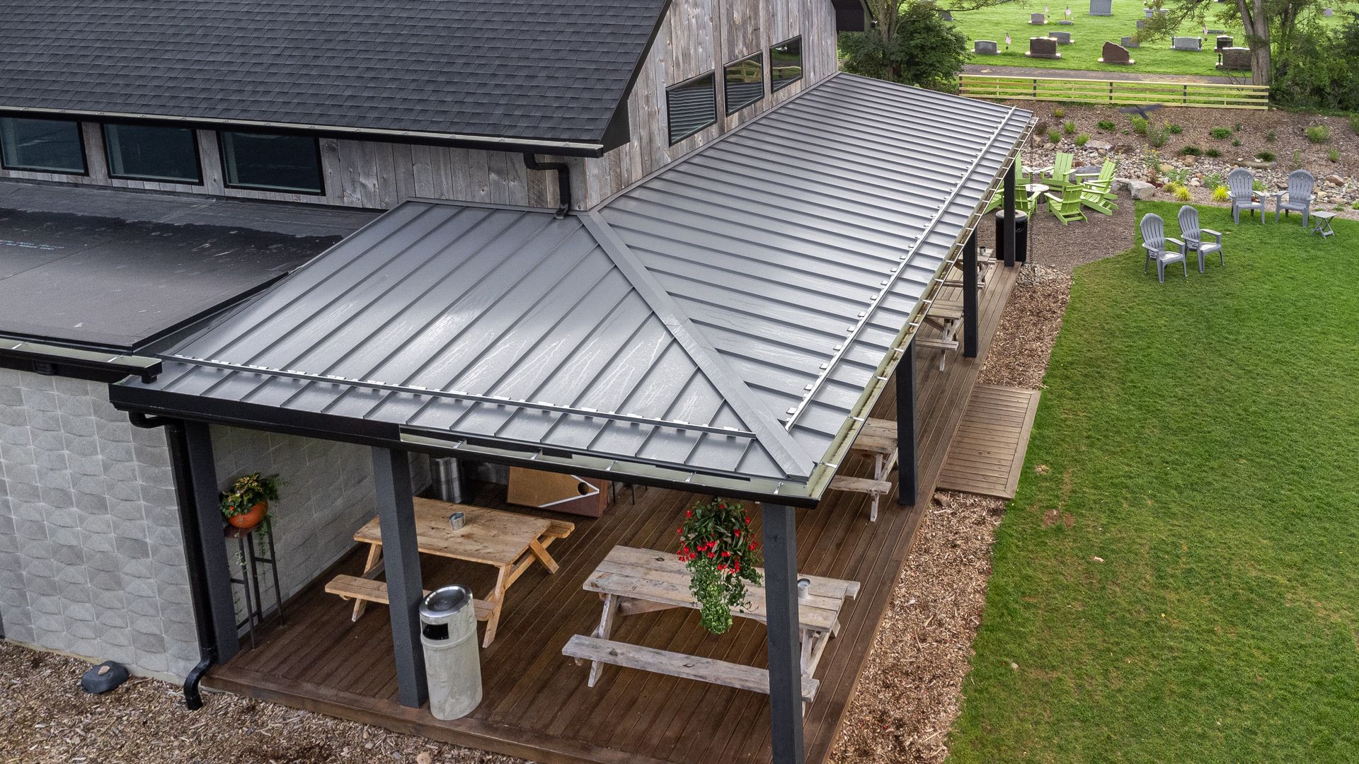 Gray-roofed outdoor patio with picnic tables, part of a building, green grass and trees in the background.