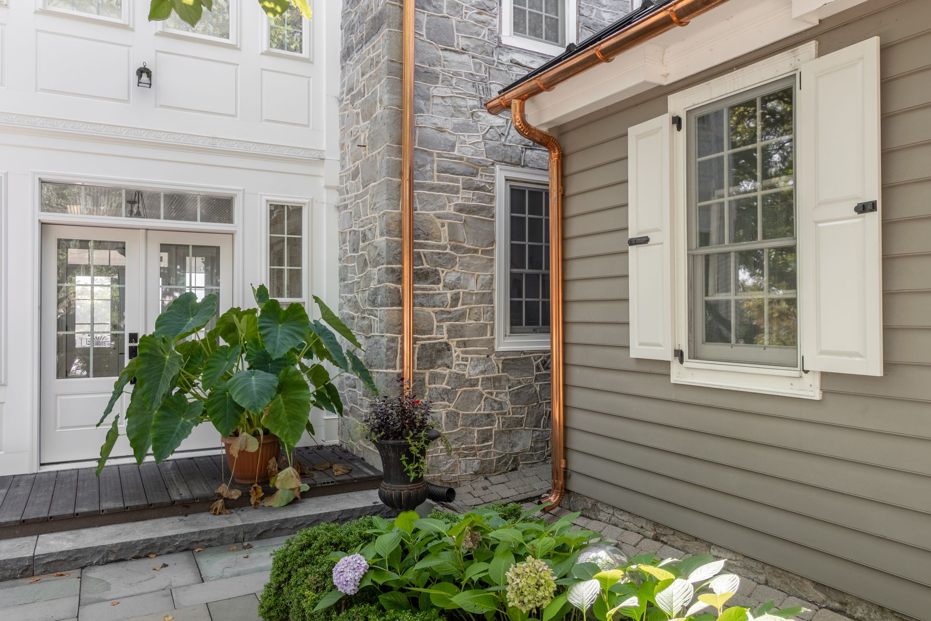 Stone and grey house exterior with copper gutters and white shutters, plants in foreground.
