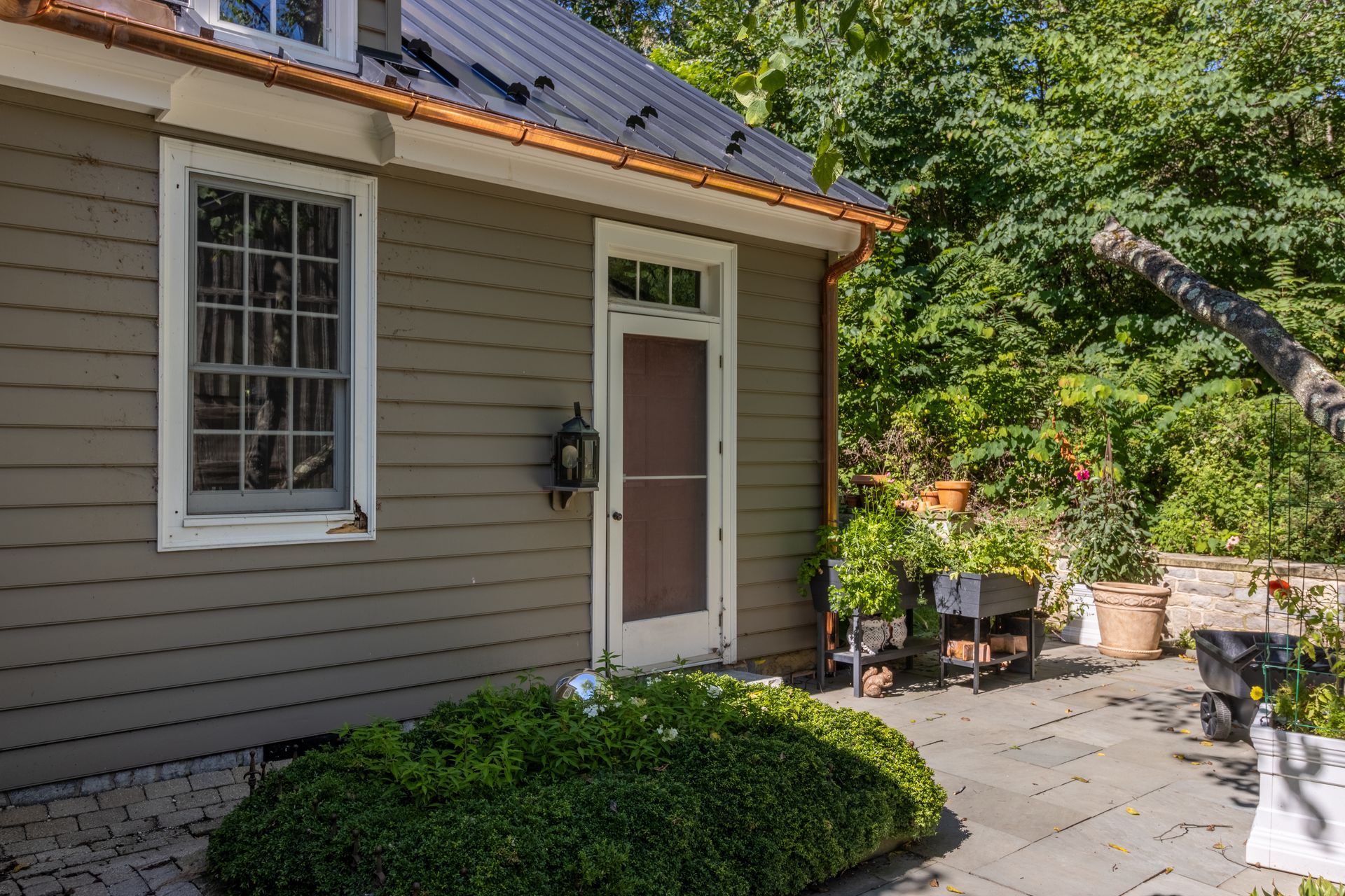 Side of a house with a window, door, and green shrubs on a patio. Garden pots and trees are visible.