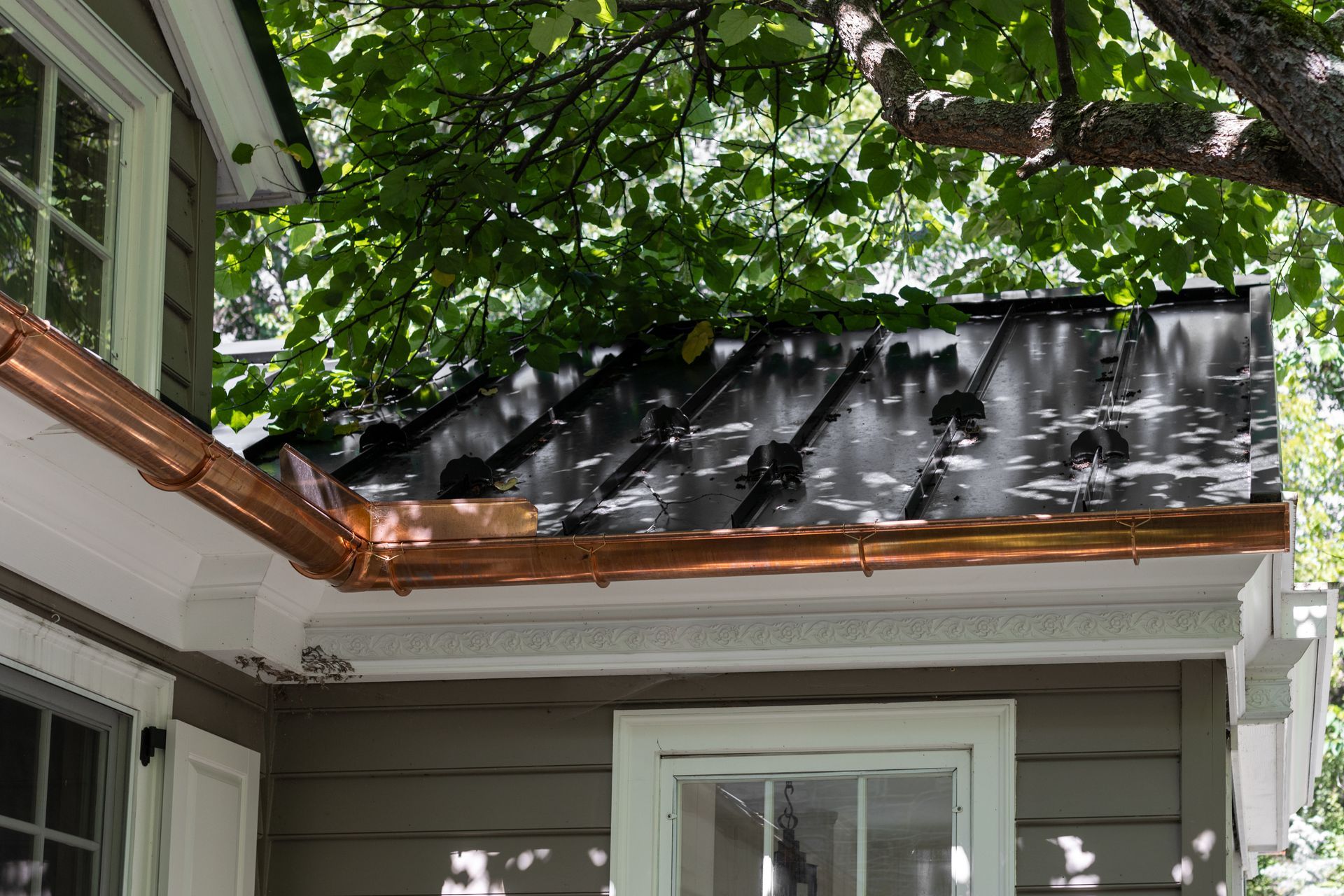Copper gutters and a dark-colored roof against a tree.  A house with white trim and windows.