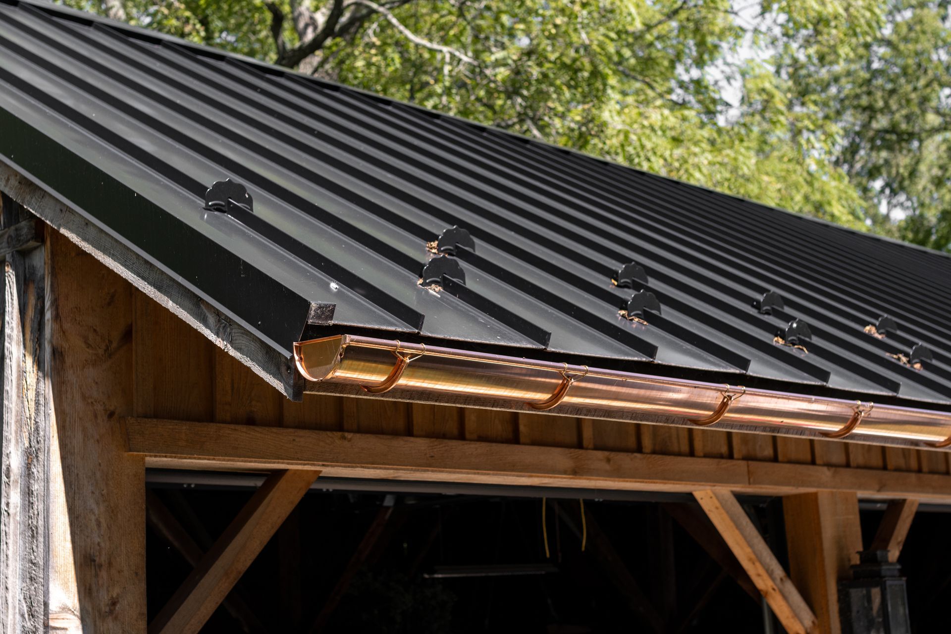 Black metal roof with copper gutters on a wooden structure, trees in the background.
