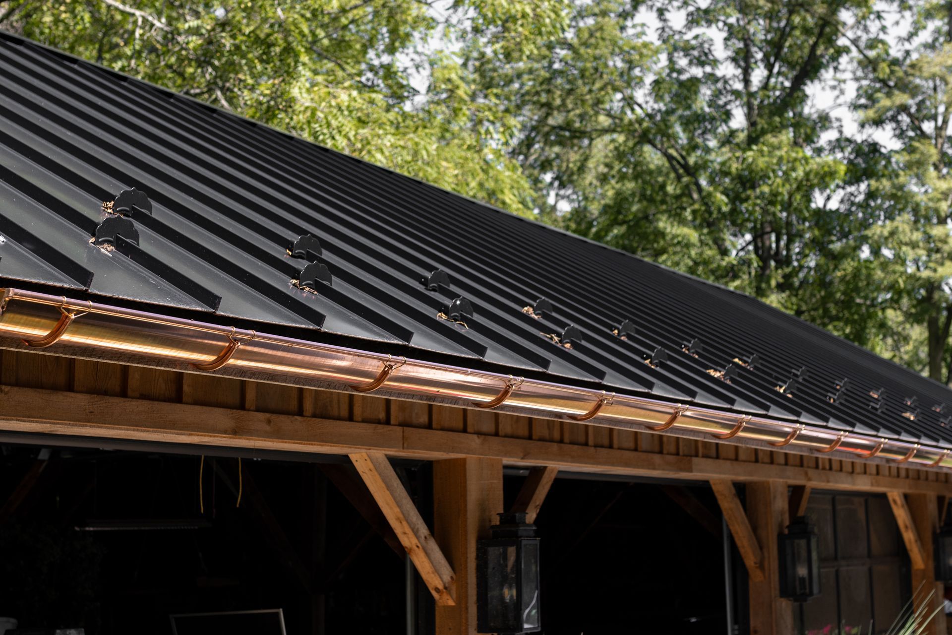 Black corrugated metal roof with copper gutters on a wooden structure. Trees in the background.