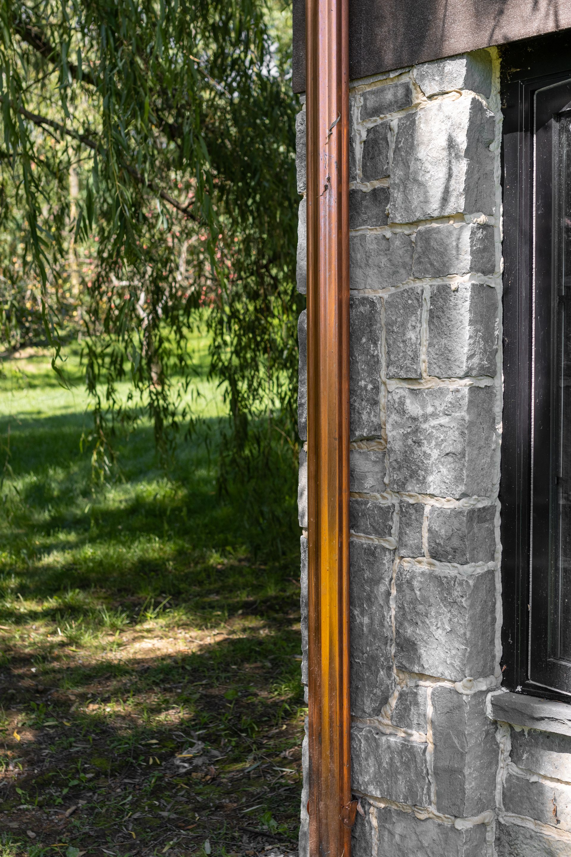 Copper rain gutter next to a weathered gray stone wall, with a window and trees in the background.