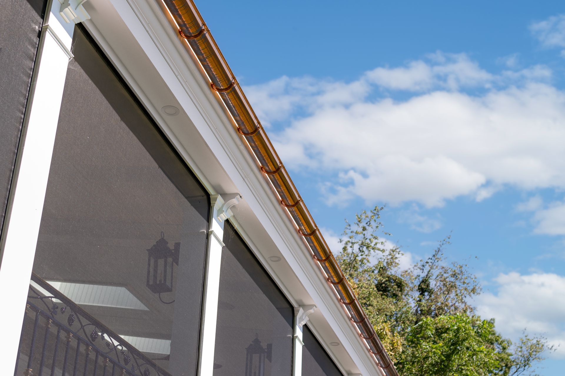 Copper gutters on a white house with dark screen windows, set against a blue sky and some greenery.