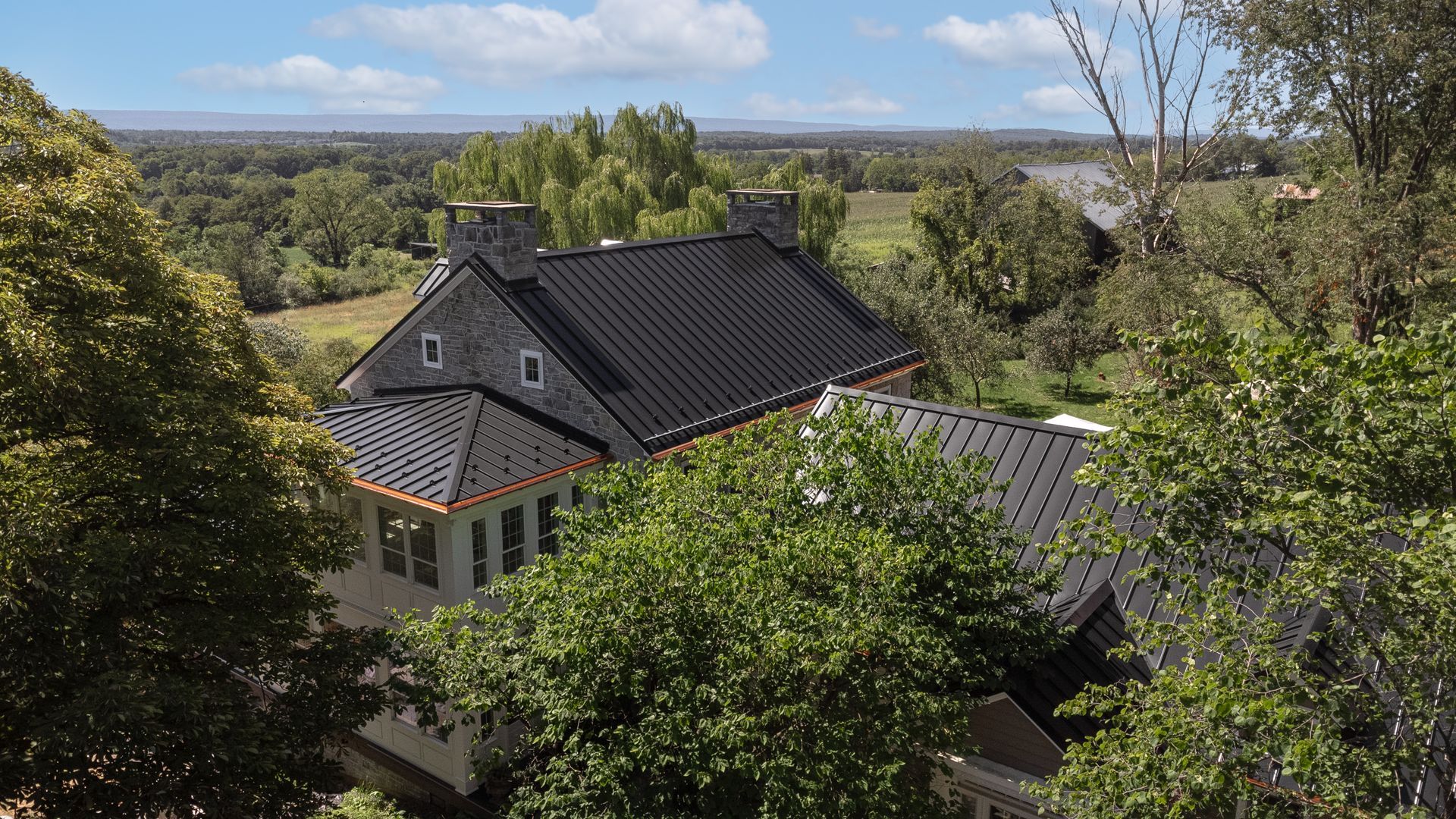 A house with a black roof, surrounded by trees, with a view of fields and hills in the distance.