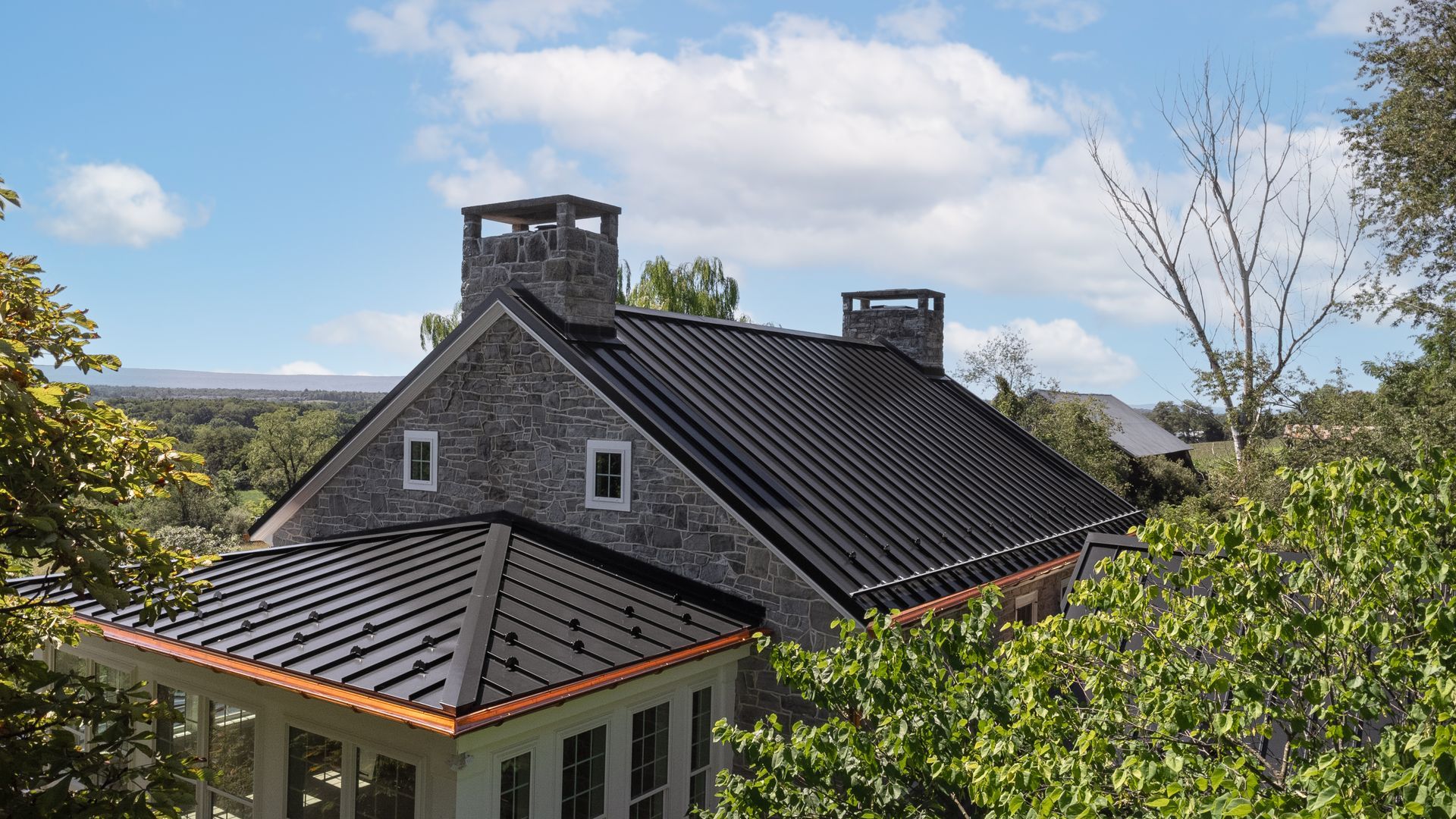Stone house with dark metal roof, two chimneys, and trees under a blue sky.