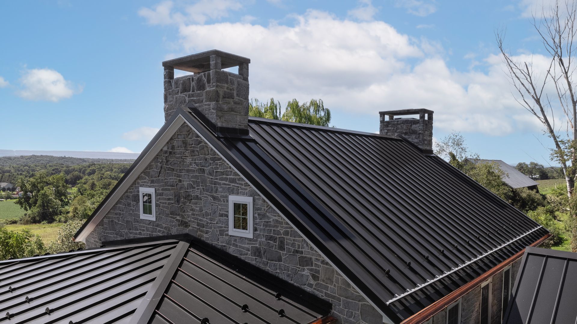 A black metal roof on a stone house with two stone chimneys, set against a blue sky.