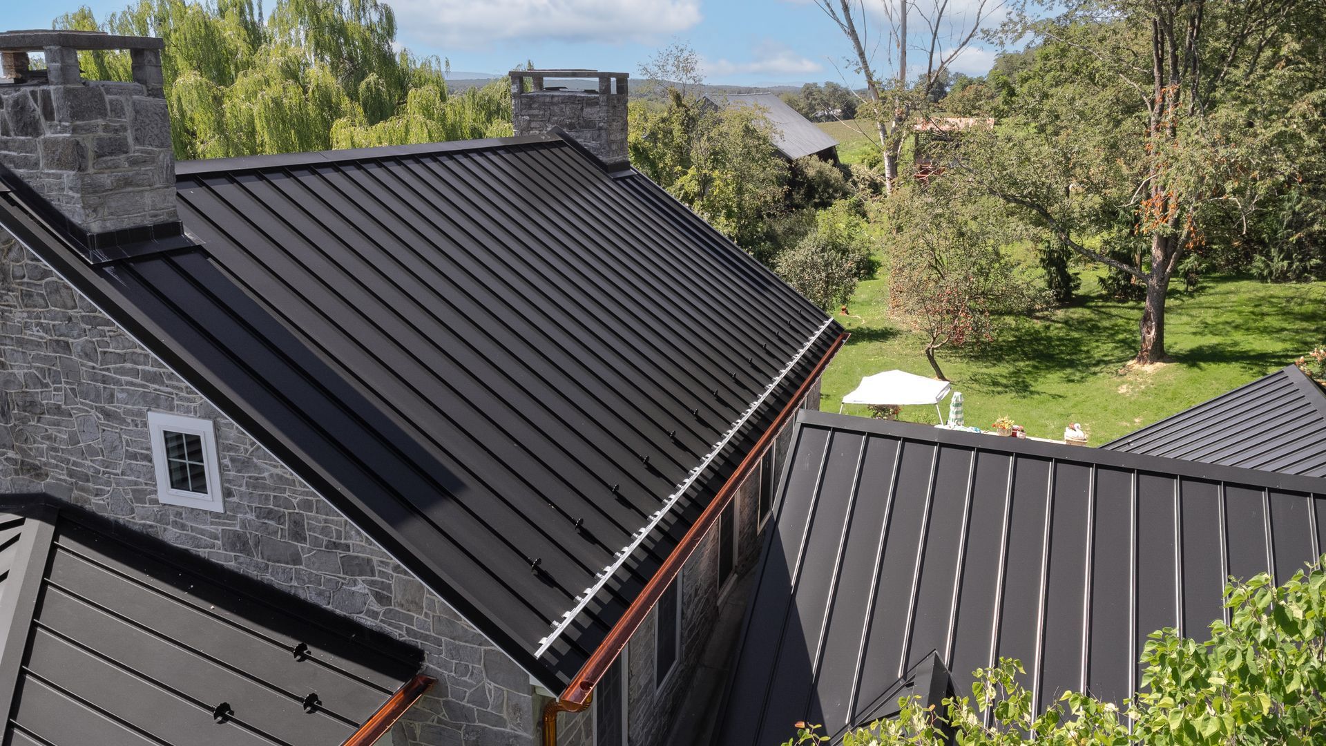 Black metal roof on a stone house with a green lawn and trees in the background.