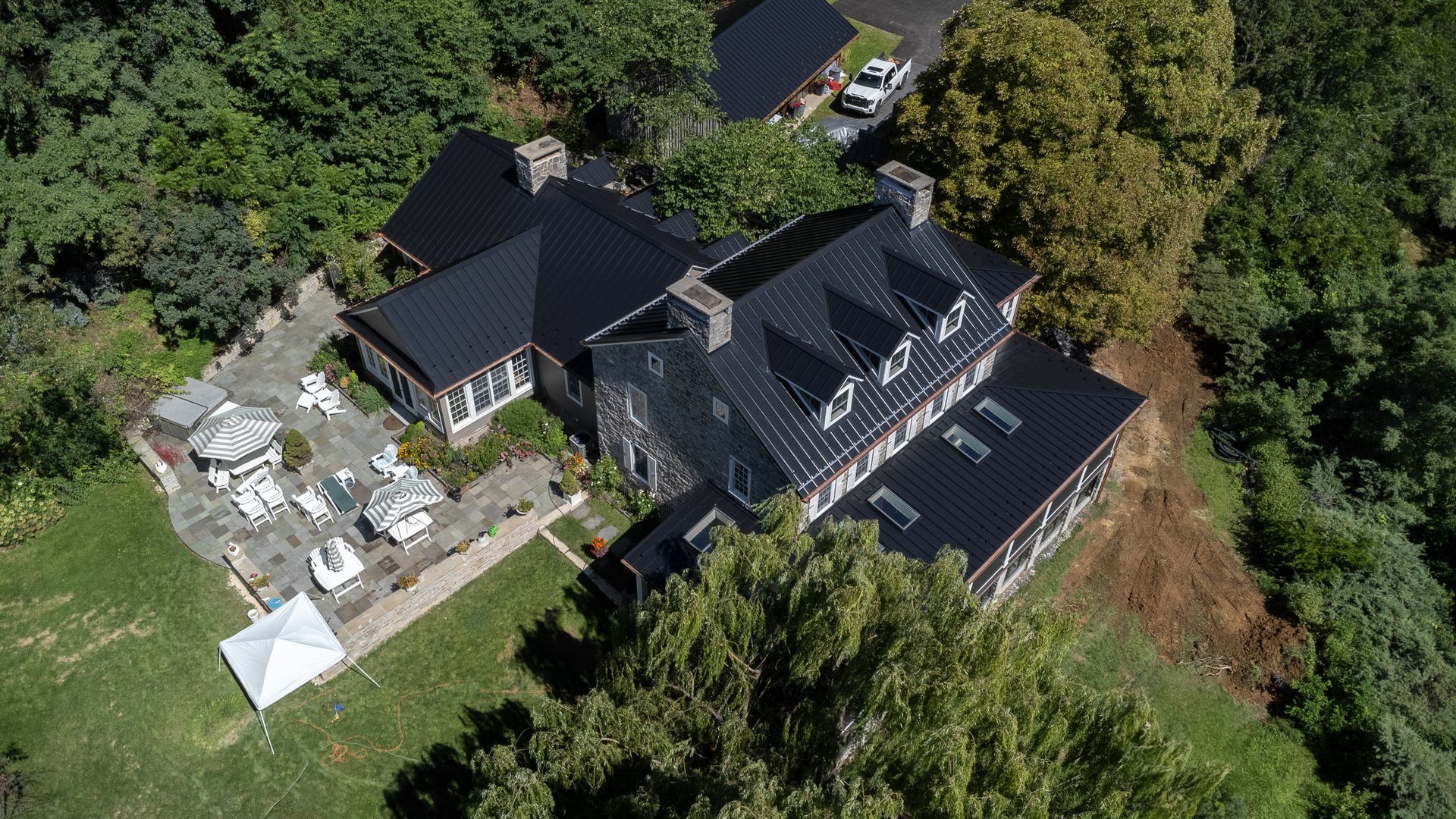 Aerial view of a large grey house with a black roof, surrounded by trees and greenery.