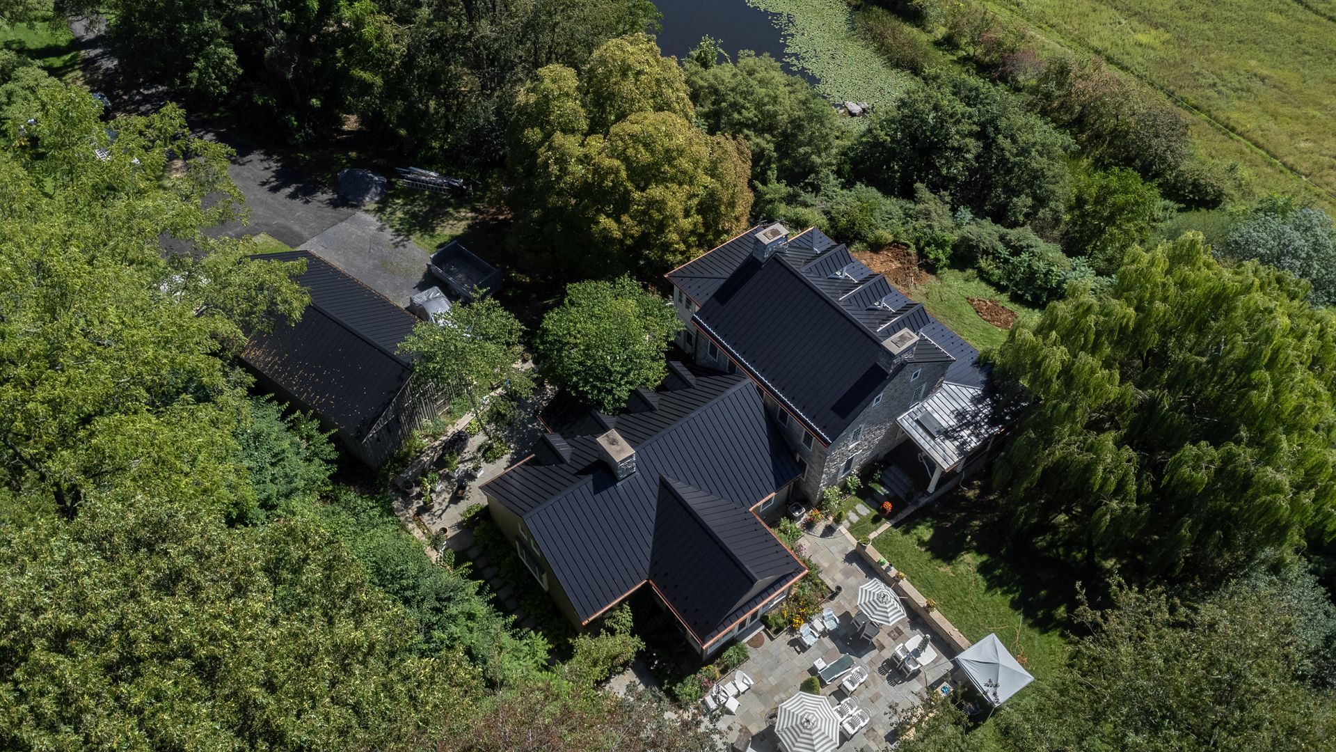Aerial view of a house with a dark roof, surrounded by trees and a lawn, in a rural setting.