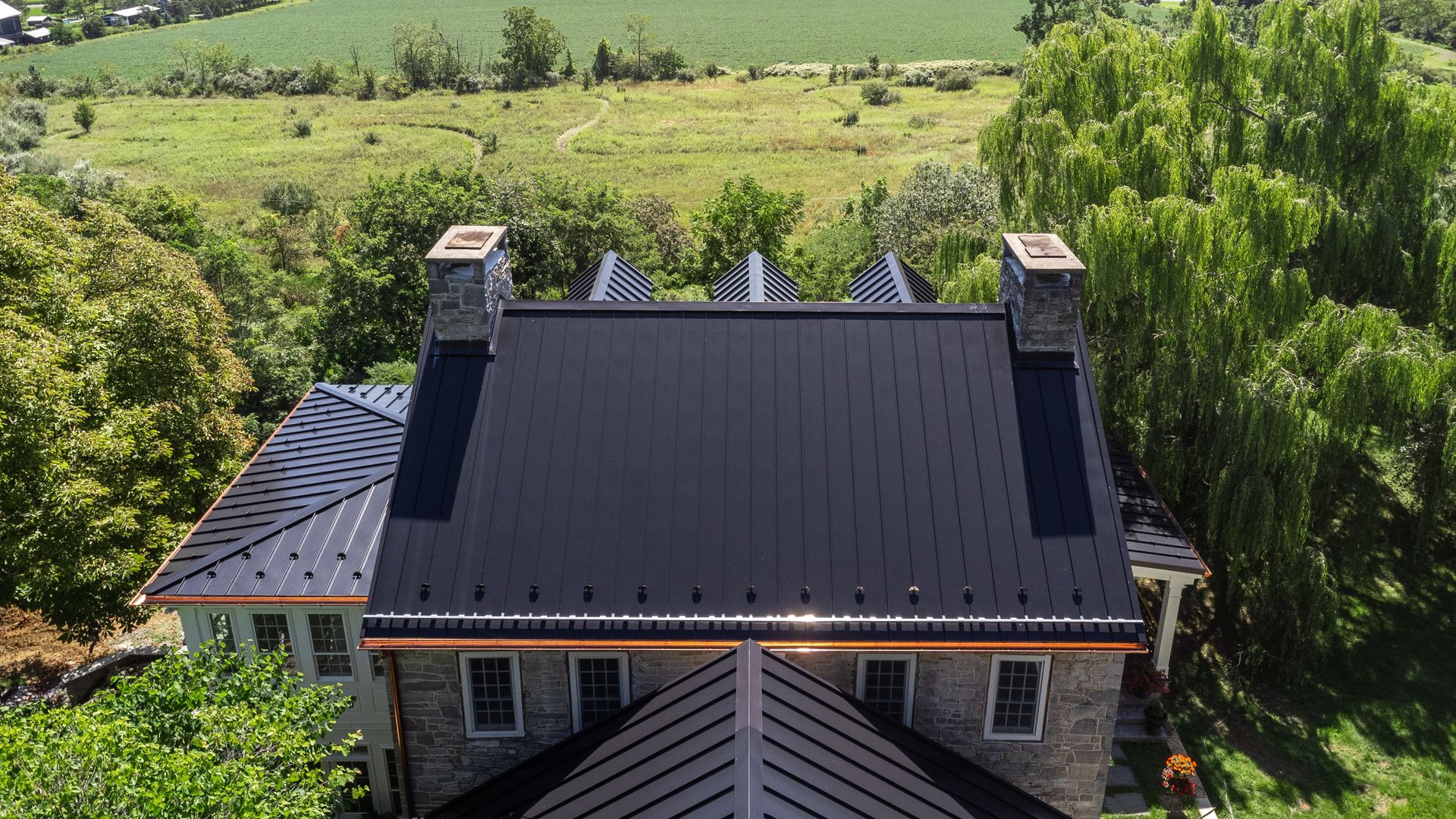 Black metal roof on a stone house with copper gutters, chimneys, and a green landscape background.