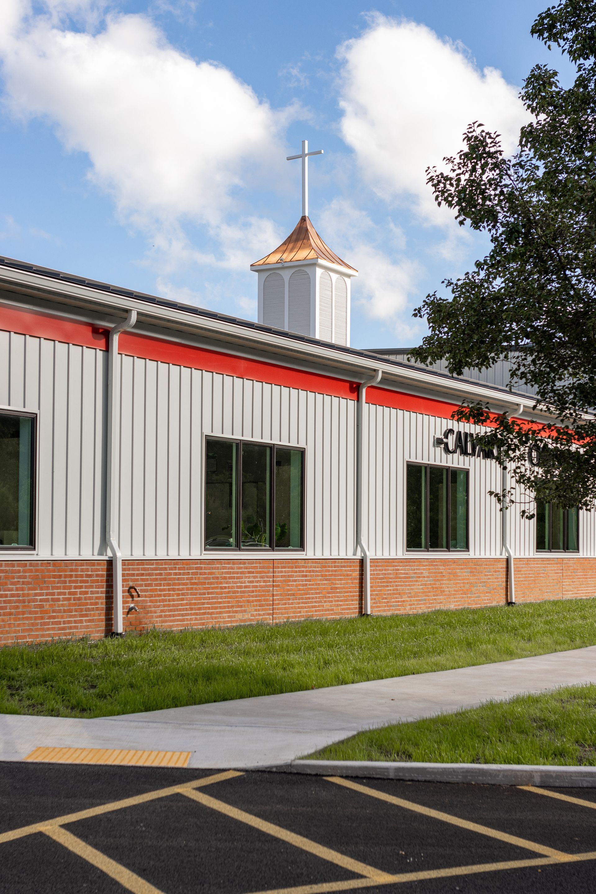 A church building with a steeple and cross, featuring a red trim, brick base, and metal siding.
