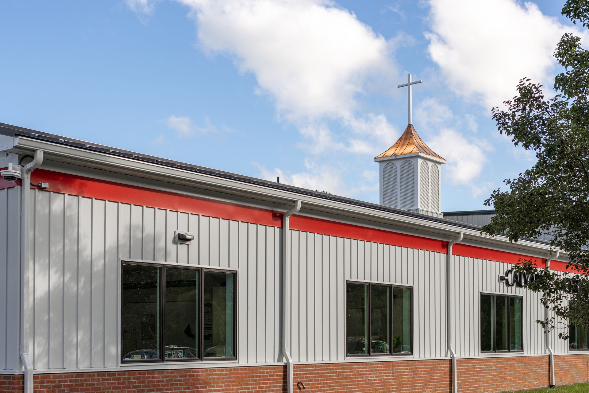 Building with metal siding, red trim, windows, and a steeple with a cross against a partly cloudy sky.