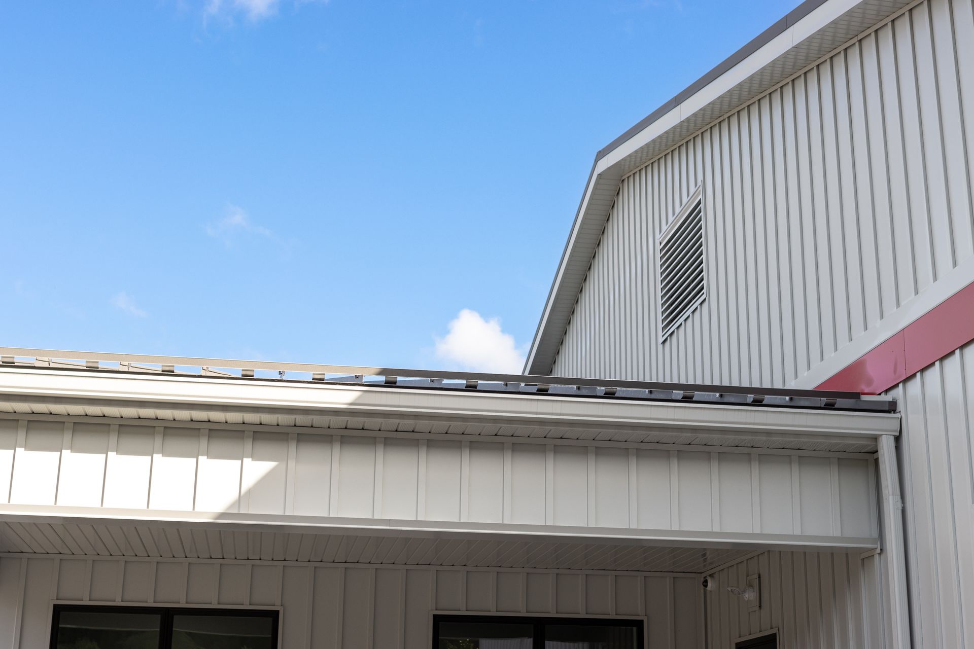 White building with a dark roof against a blue sky; includes a ventilation grate and some windows.
