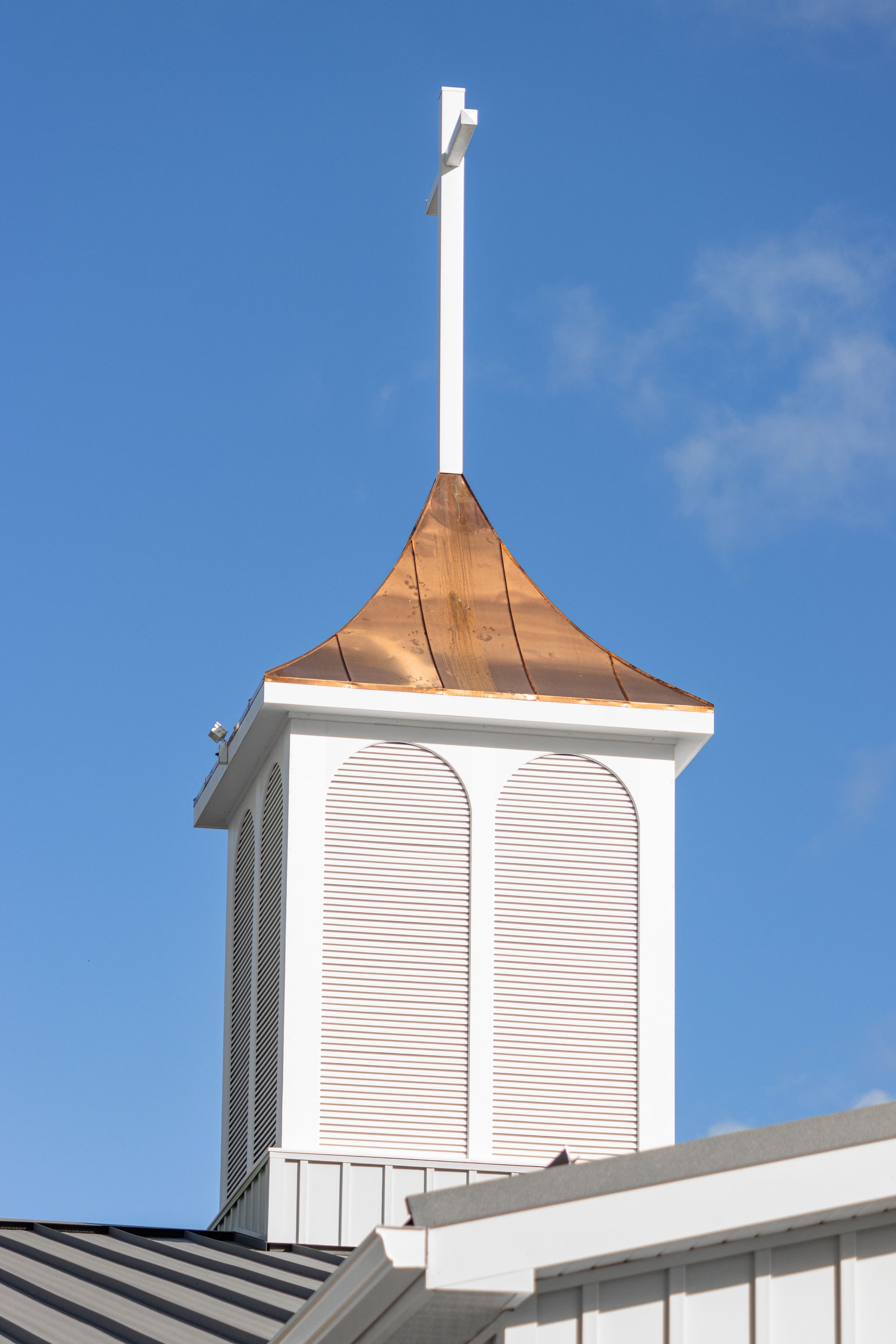 White steeple with copper roof, topped with a cross, against a blue sky.