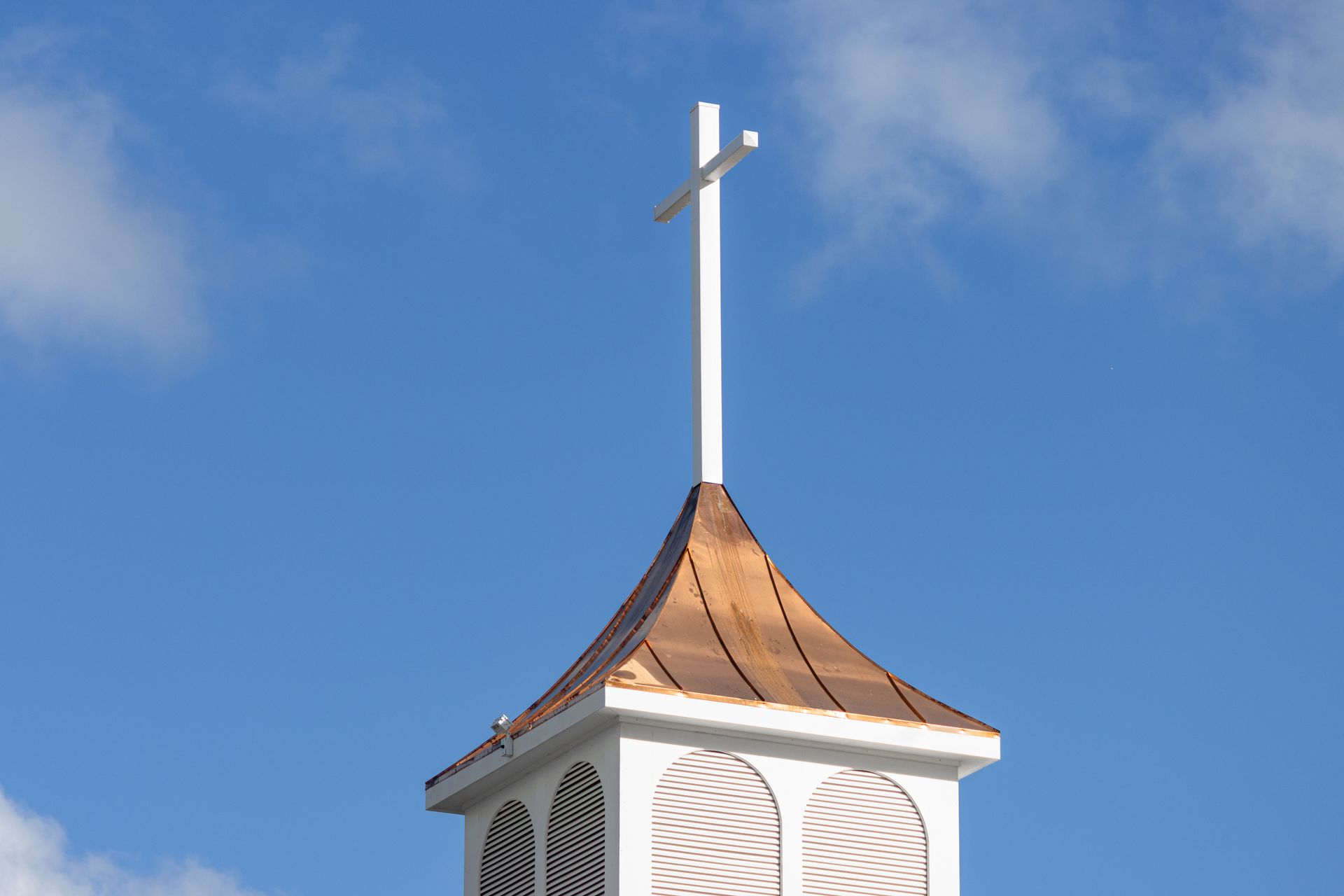 White cross atop a copper-roofed church steeple against a blue sky.