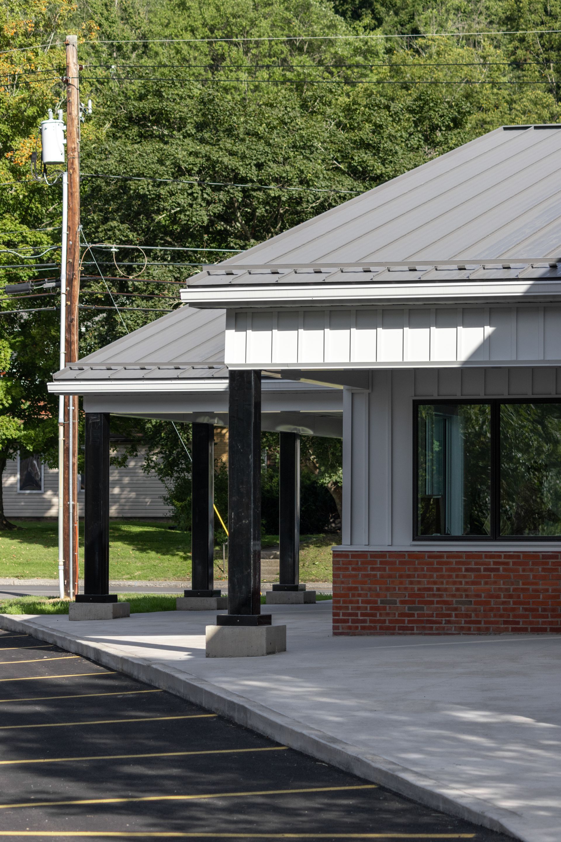 Gray building with black columns, metal roof, and brick base; outdoors, trees in background.