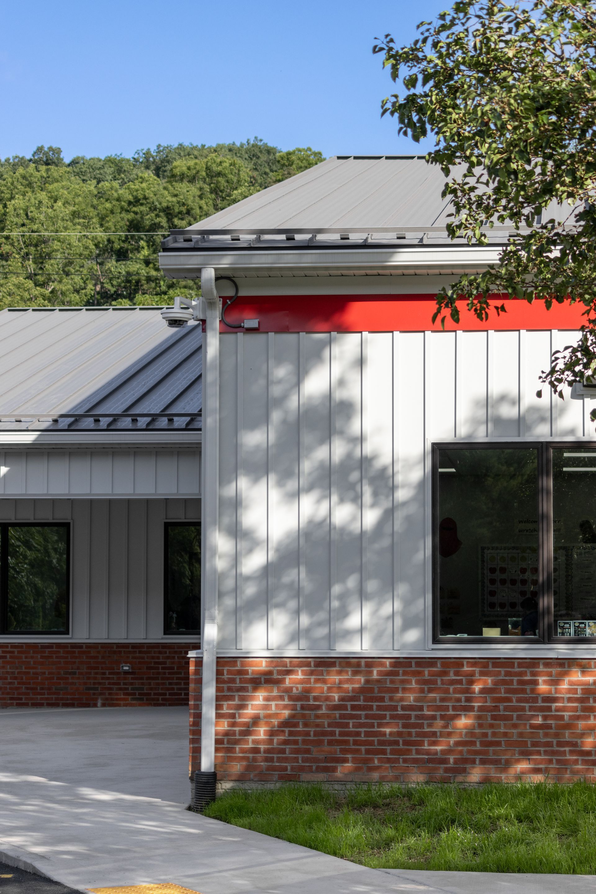 White building with red accents and a brick base, with a gray metal roof and trees in the background.