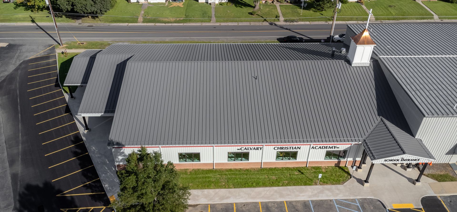 Aerial view of a building with a gray metal roof and a white steeple. Parking lot and street visible.