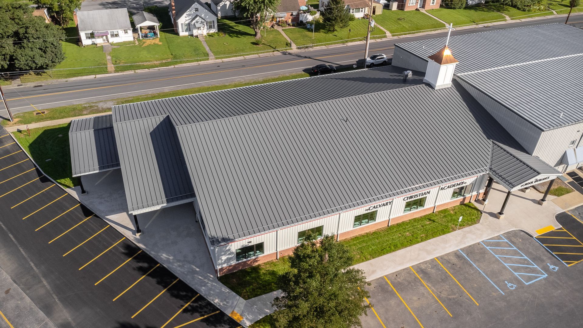 Aerial view of a gray-roofed church with a parking lot and street in front, and houses in the background.