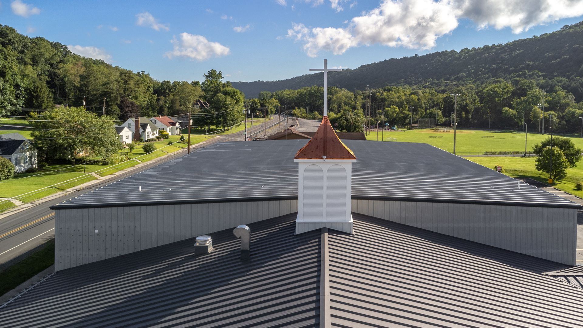 Church roof with steeple and cross, against a backdrop of a valley and trees.