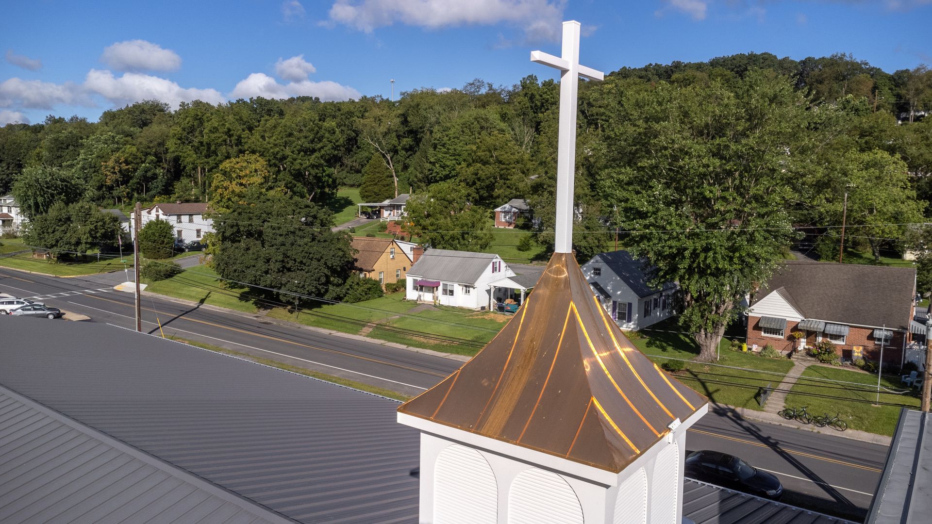 Church steeple with a cross against a backdrop of trees and houses.
