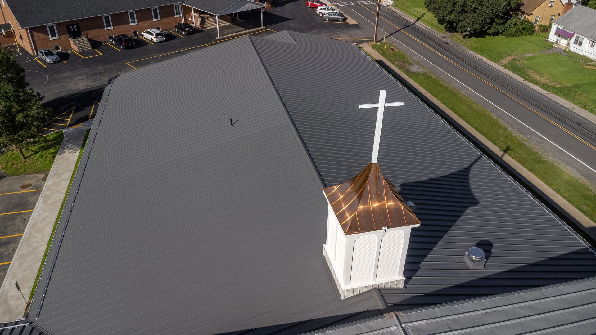 Church roof with steeple and cross; dark gray metal roofing, roadside view.