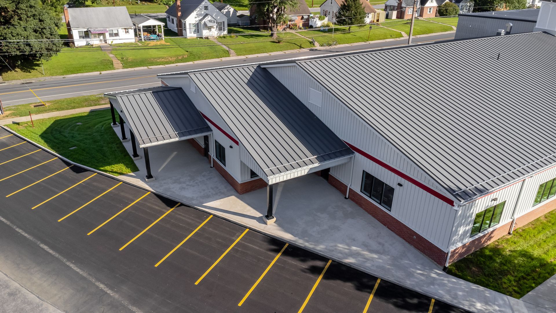 Exterior view of a modern building with parking lot. Grey metal roof, red trim, green grass, and nearby residential houses.