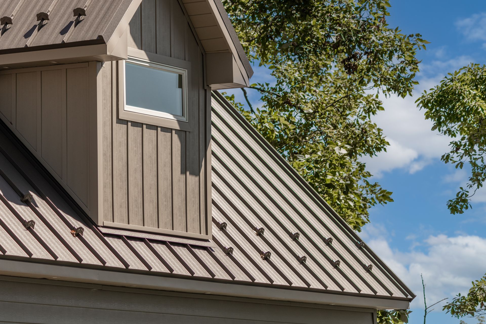 Tan metal roof on a building with a small window and siding, against a blue sky.