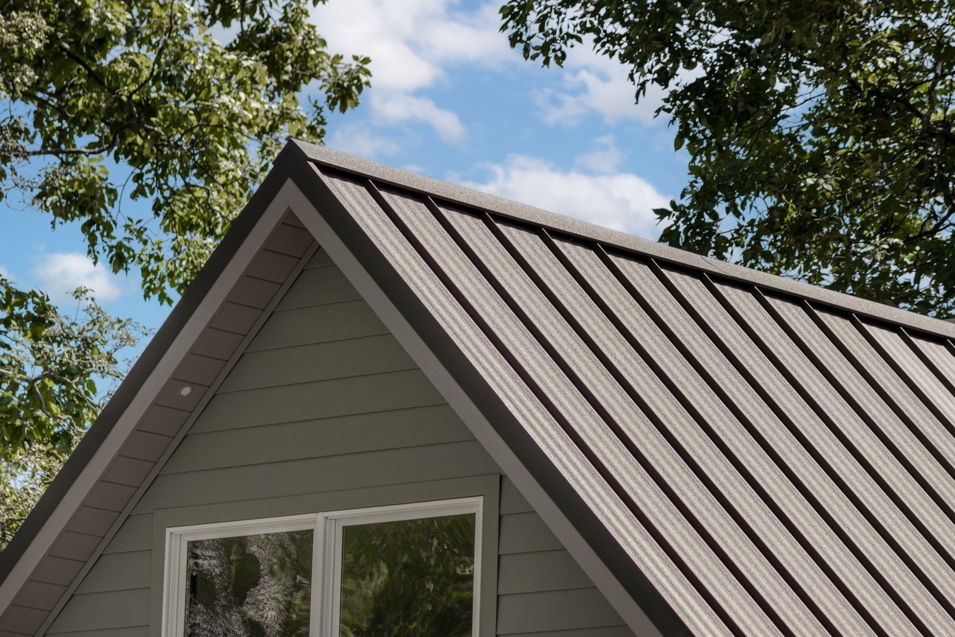 Gray metal roof on a gray house with a window, against a blue sky with trees.