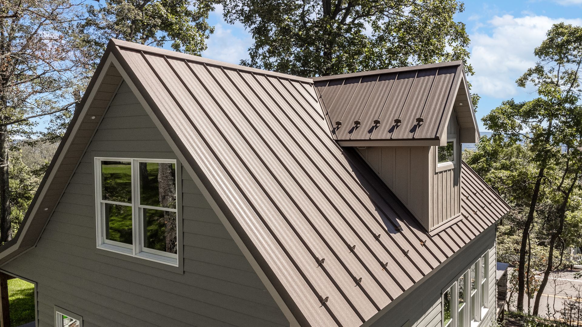 Brown metal roof on a gray house with windows, set among trees under a blue sky.
