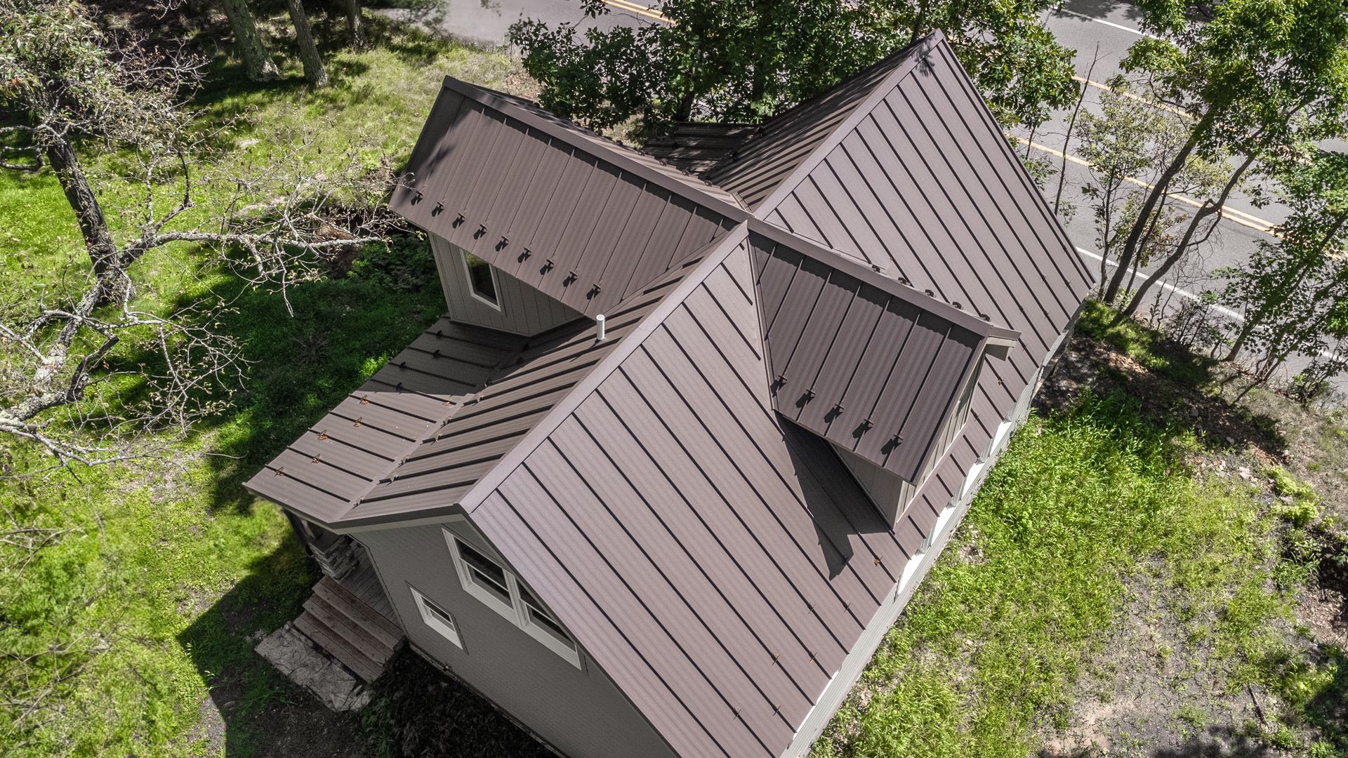 Brown metal roof on a gray house with green grass and trees surrounding it.