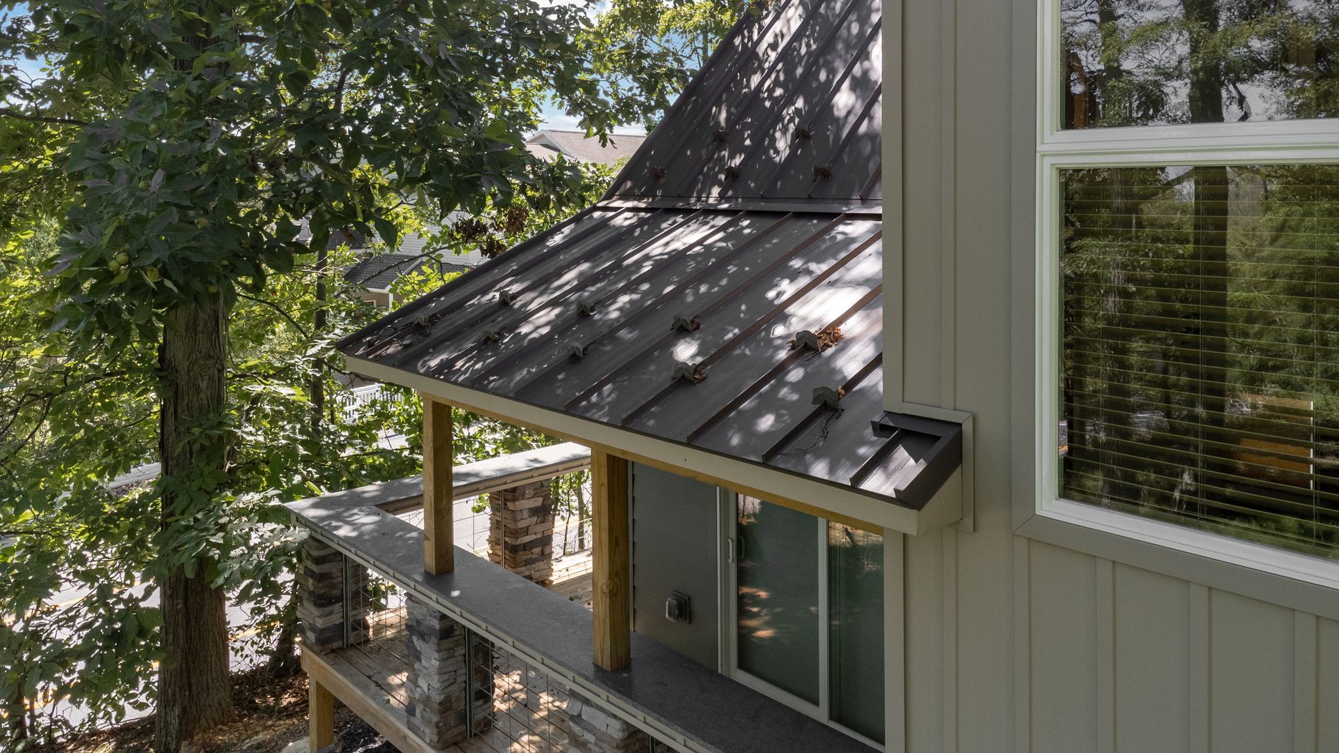 A modern house with a dark metal roof and stone pillars, surrounded by trees.