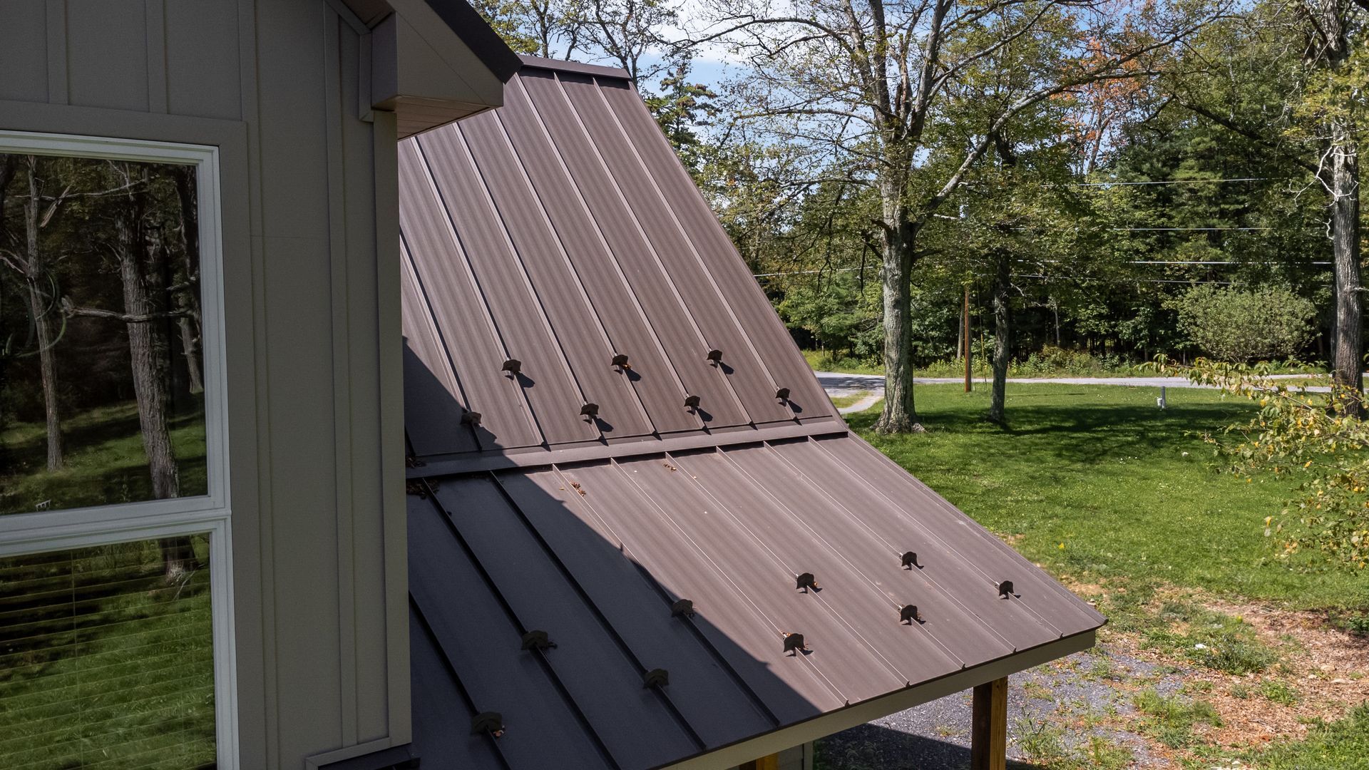 Brown metal roof with dark fasteners on a small building, green yard and trees in background.